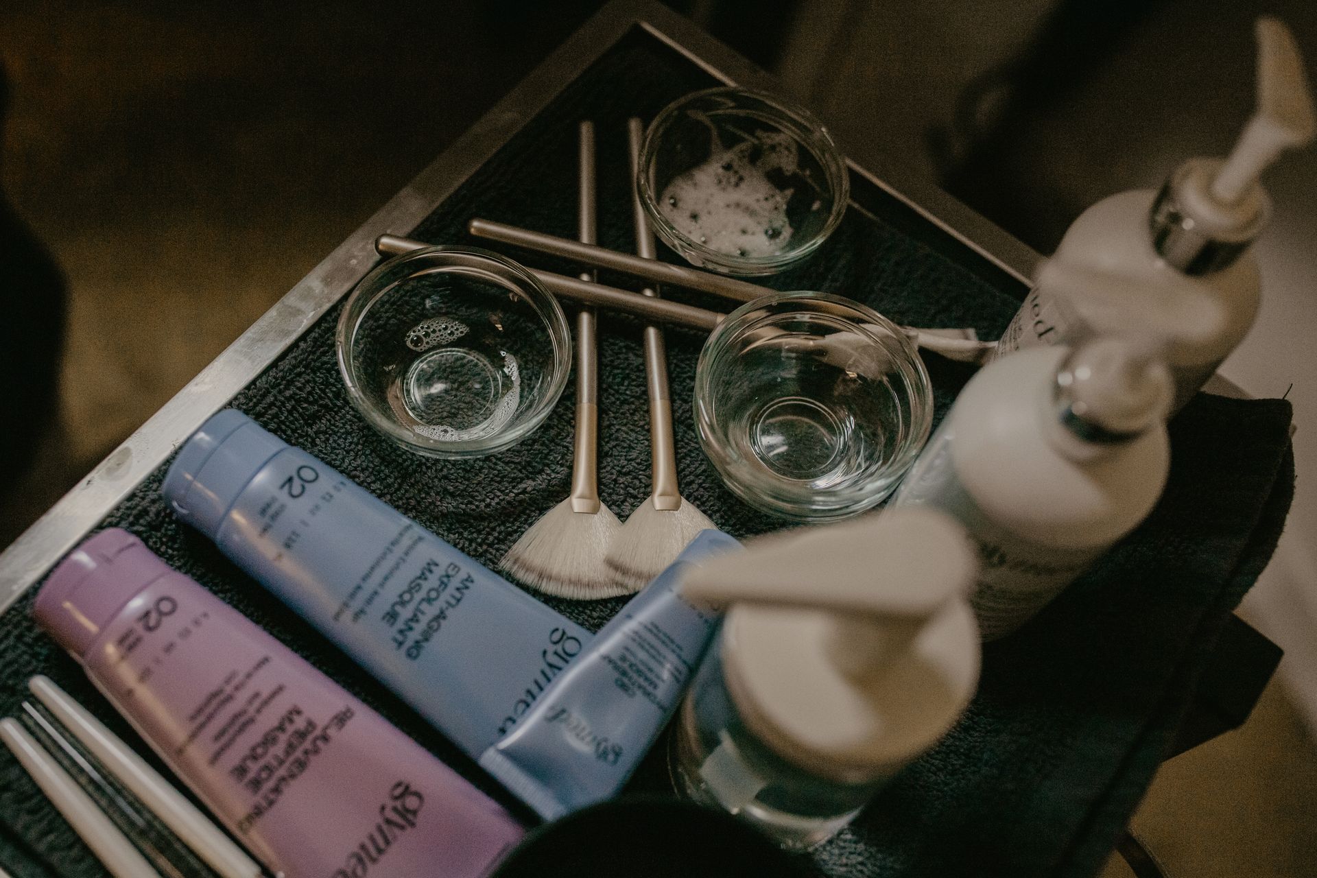 A tray holds skincare products, including tubes of purple and blue cream, small bowls, brushes, and pump bottles.