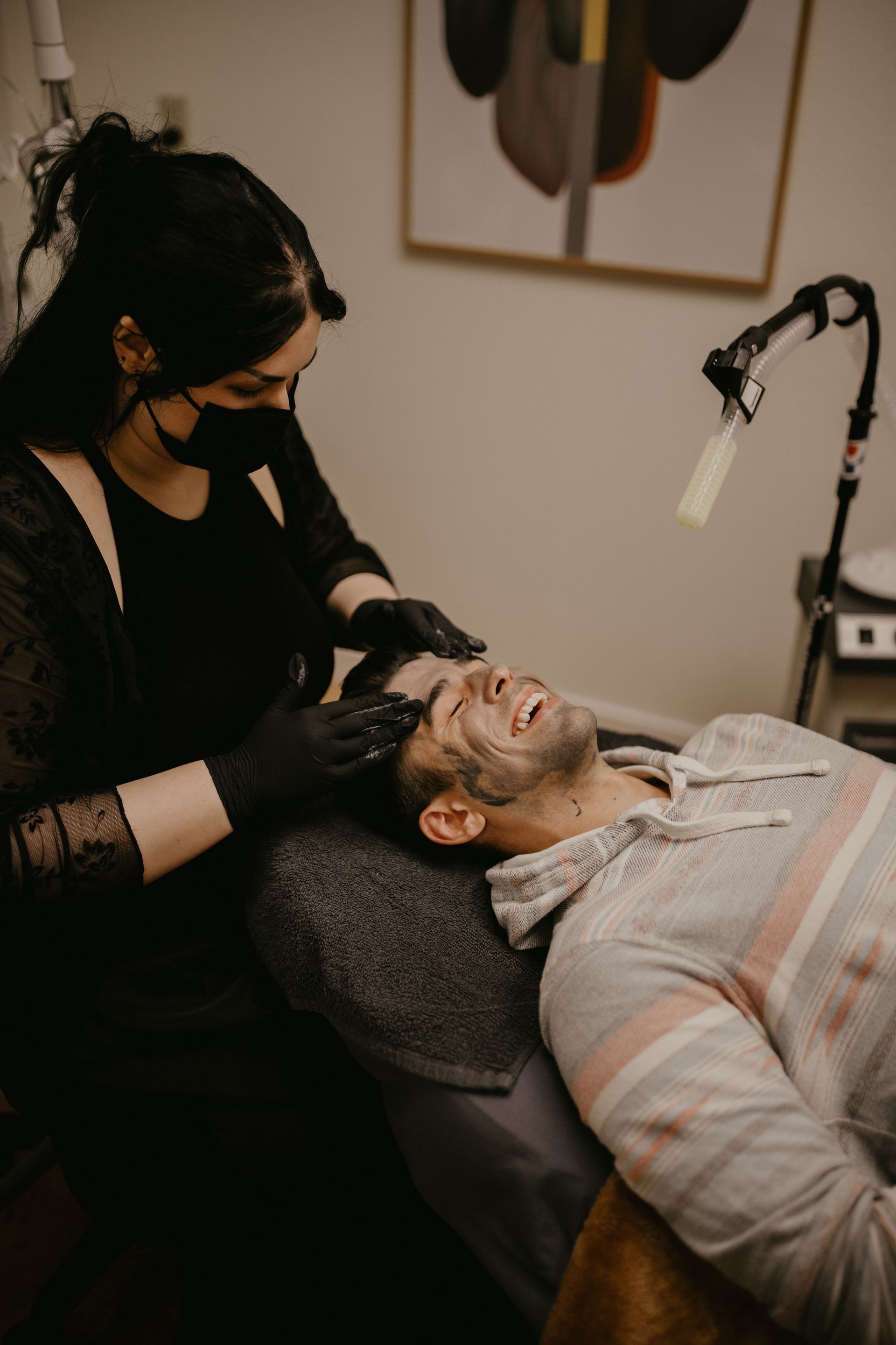Woman with mask, wearing black gloves, working on a person's face lying on a chair in a room.