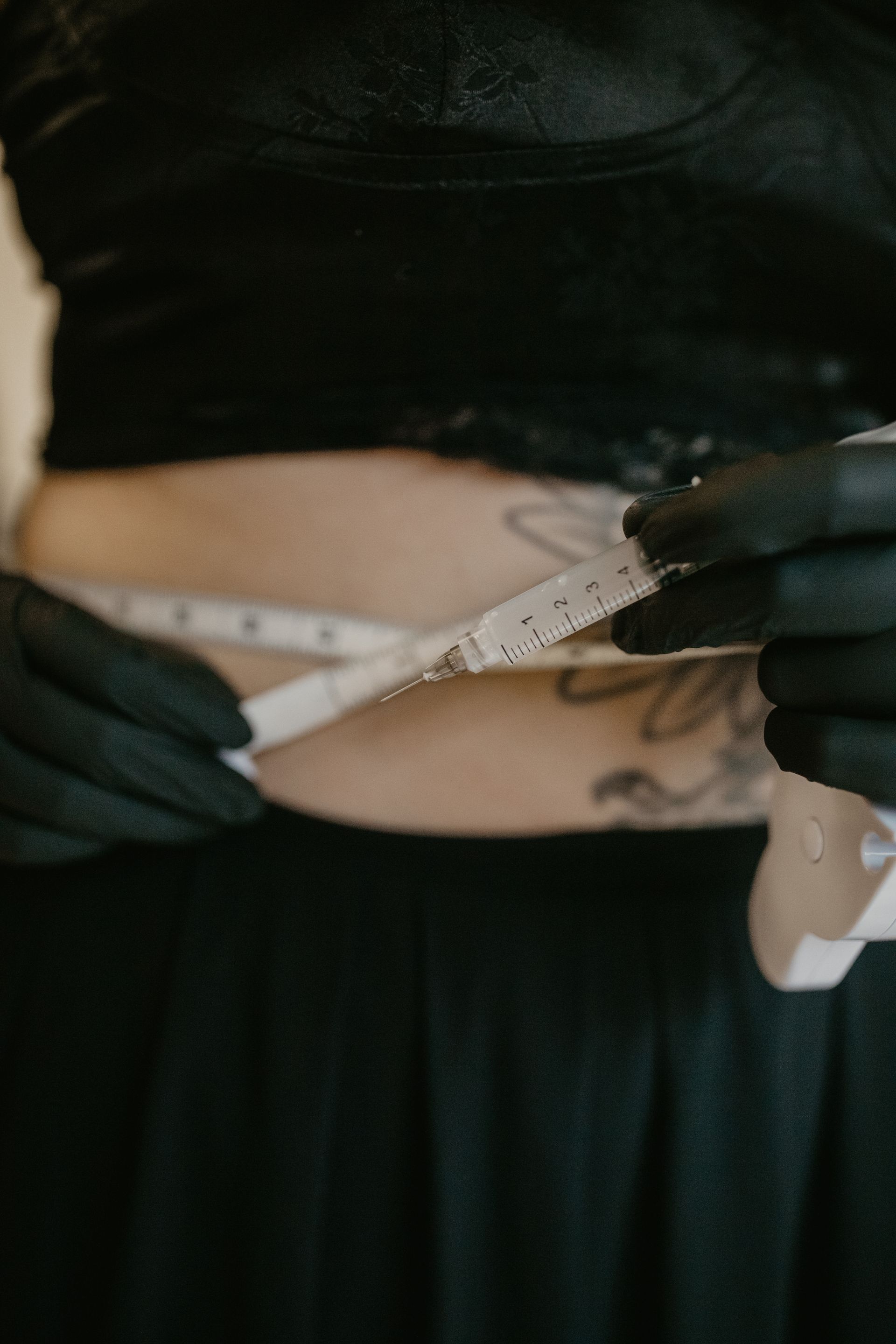 Person in black clothing measures their waist with a tape measure; black gloves are worn.