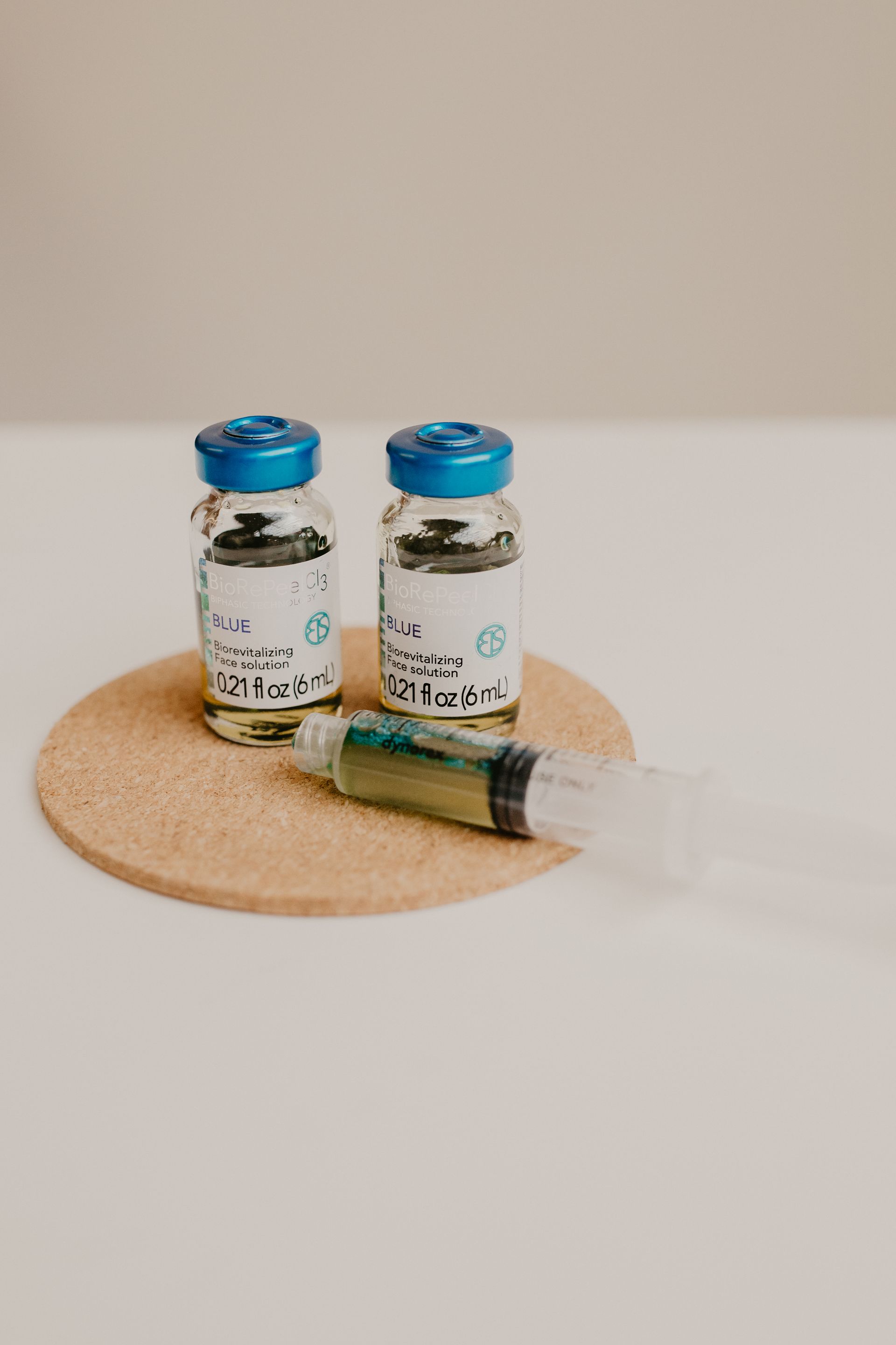 Two vaccine vials and a syringe filled with green liquid on a cork coaster, white background.
