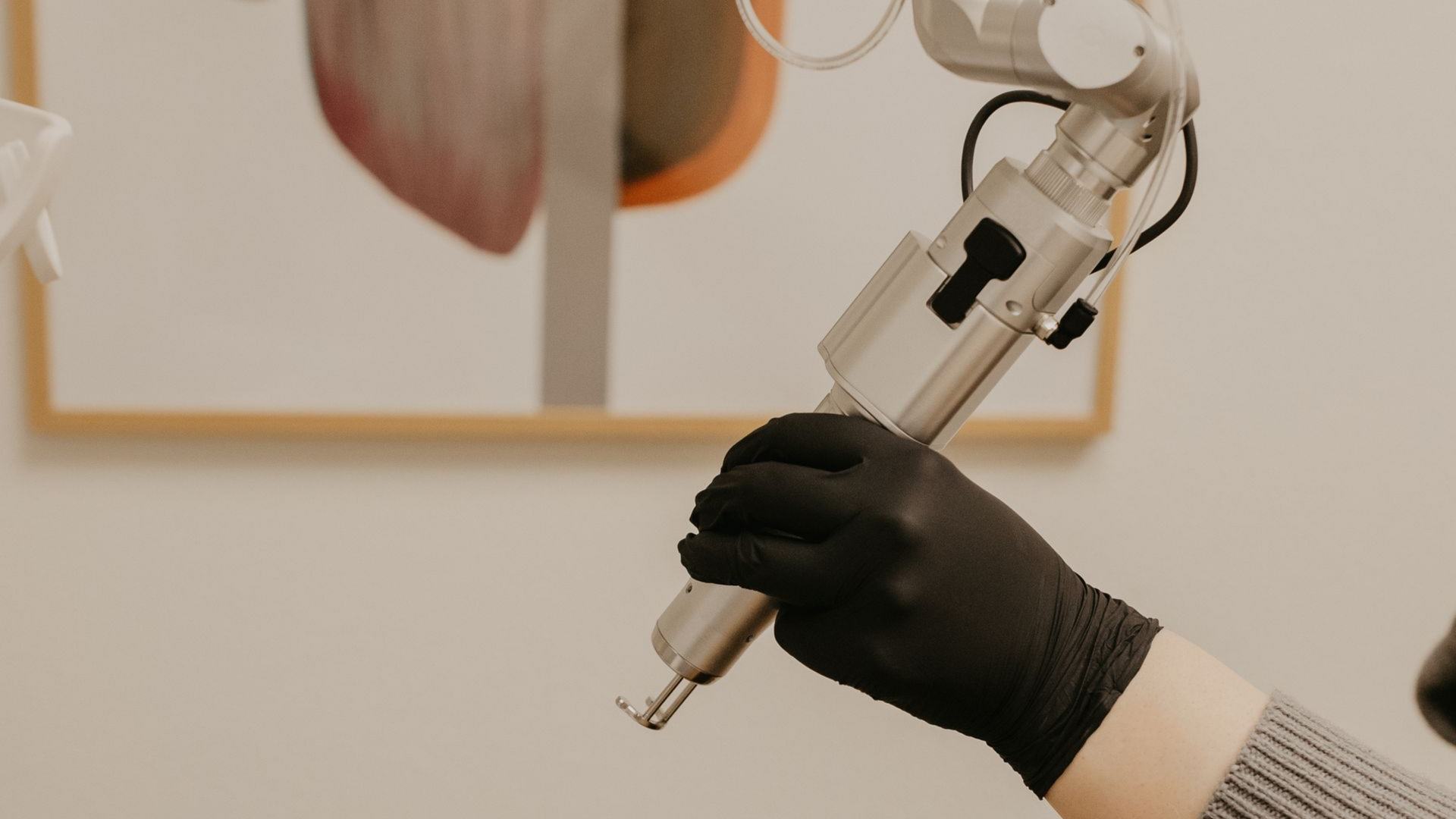 Hand in black glove holding a medical laser device, against a neutral-toned wall with art.