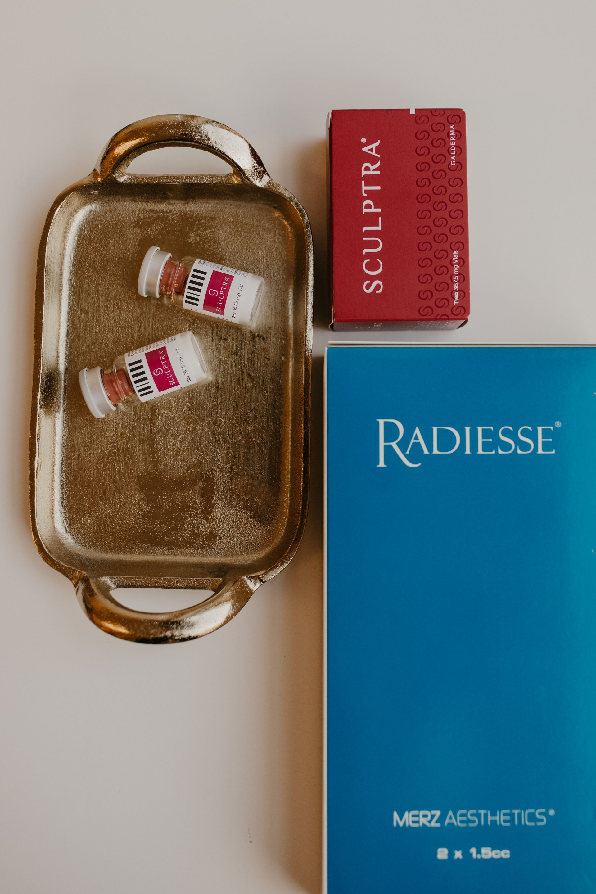 Gold tray with vials and boxes of Sculptra and Radiesse dermal fillers.