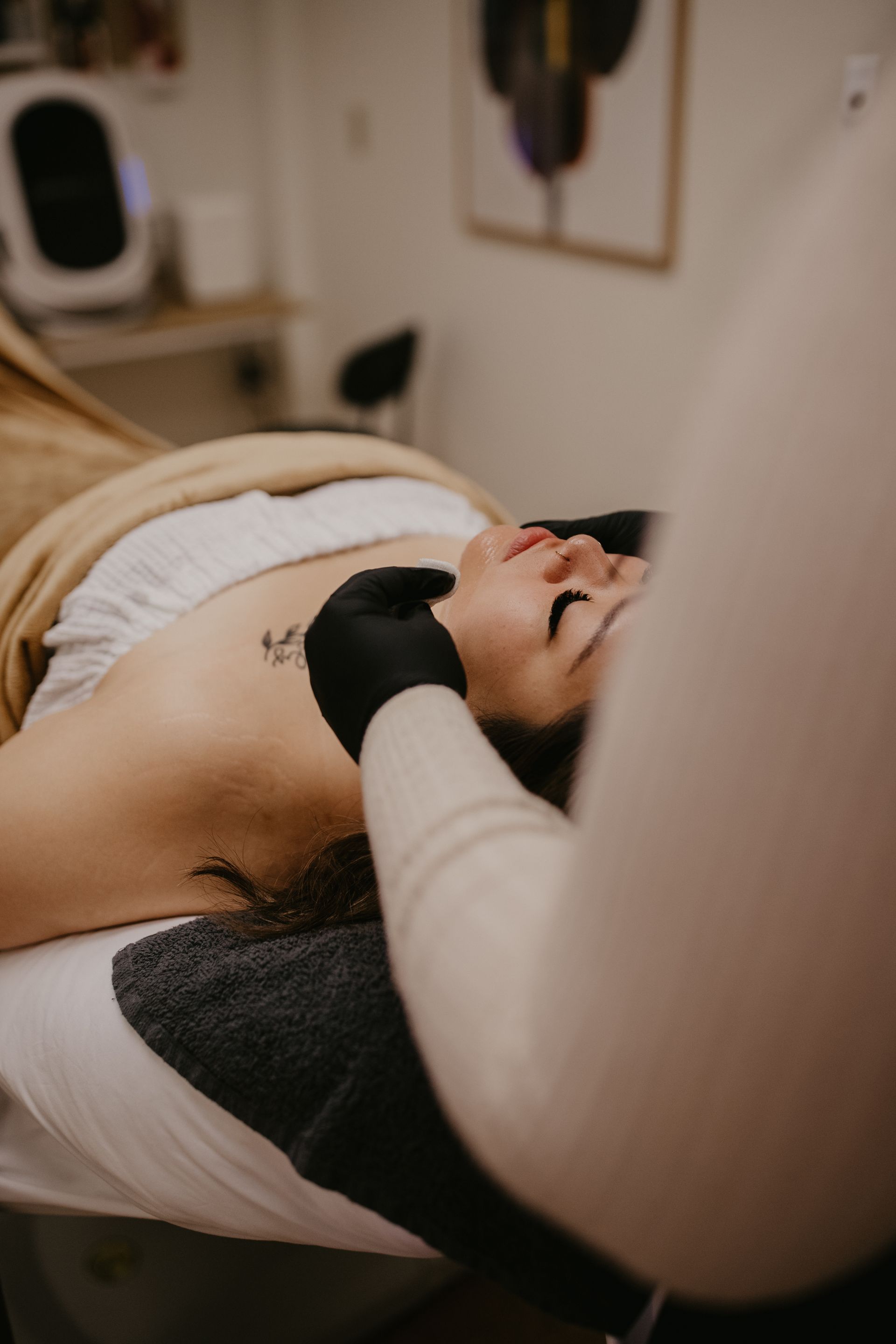 Person receiving facial treatment on a massage table. Esthetician's gloved hands on the client's face, indoor setting.