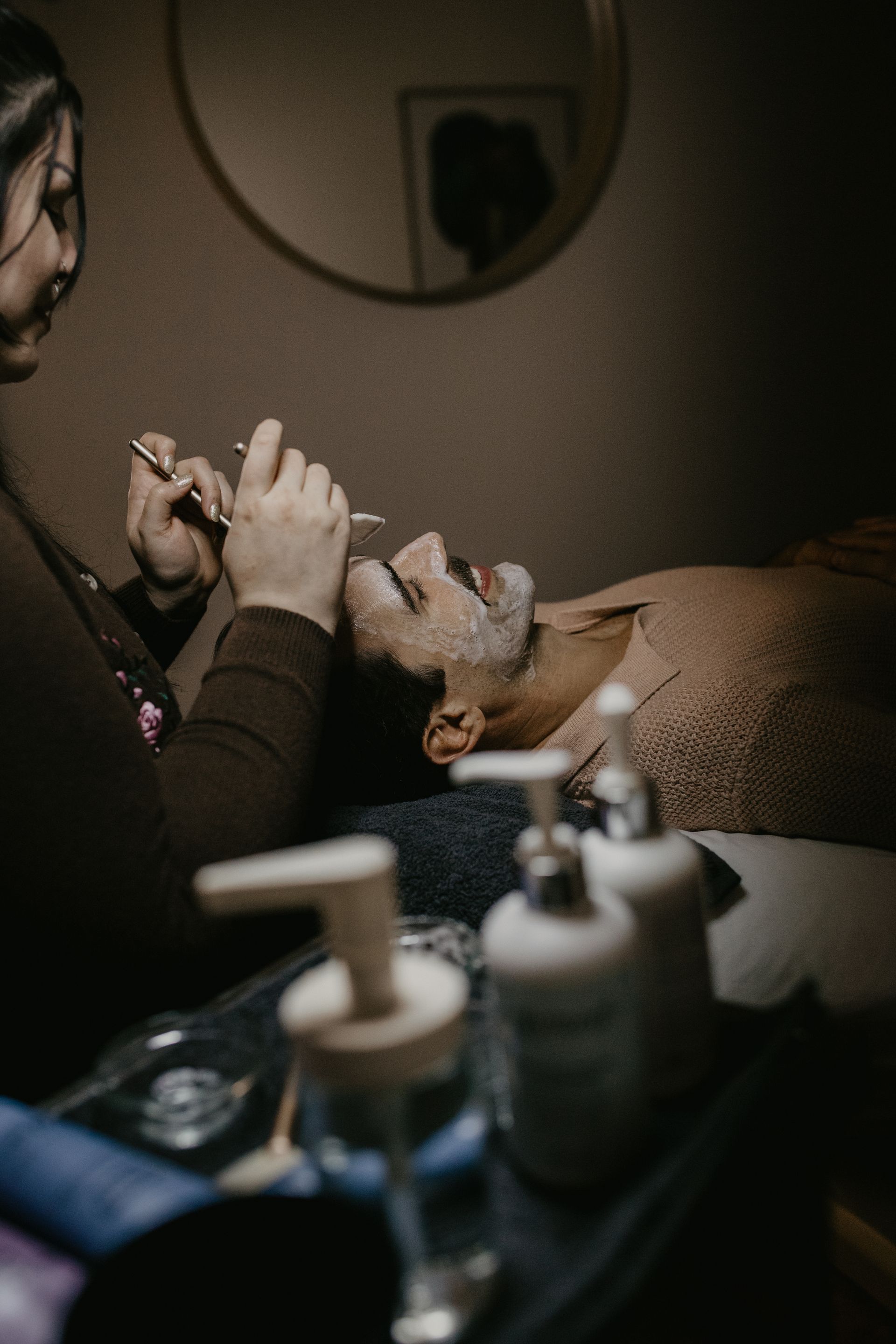Person receiving facial treatment; therapist applying products. Dark room, neutral colors.