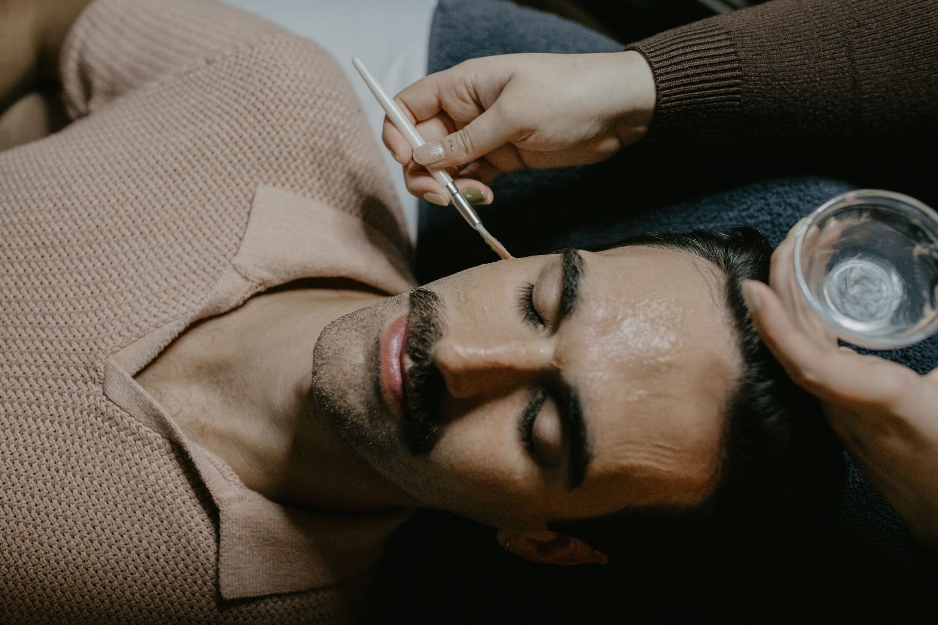 Man receiving facial treatment, being touched with a brush, bowl of liquid nearby.