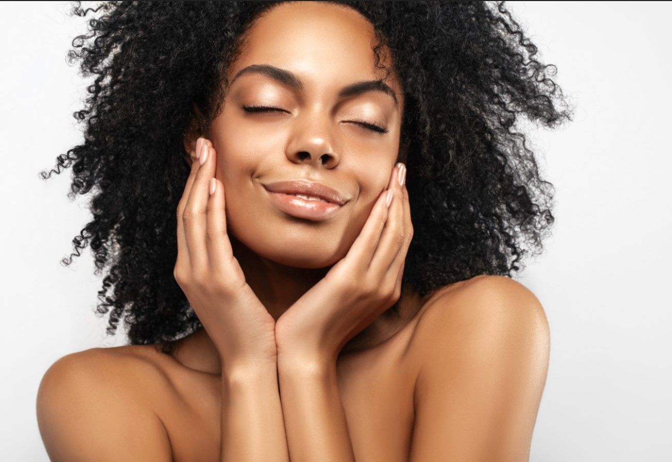 A woman with curly hair is smiling and touching her face with her hands.