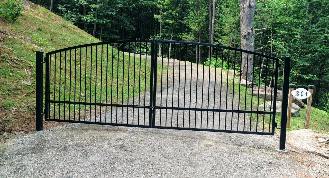 Black metal driveway gate at a gravel entrance, with trees and a mailbox on the right.