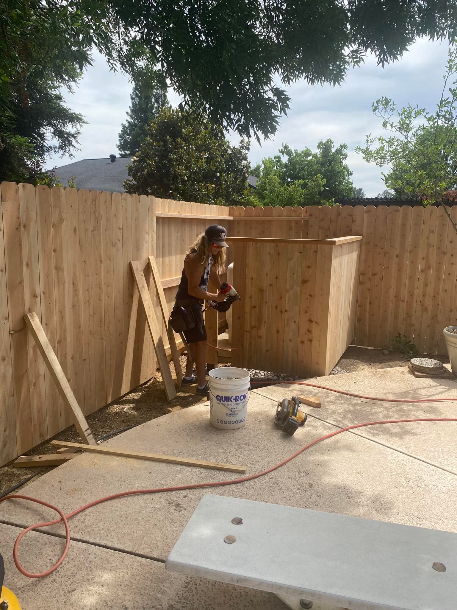 Person in a hat working beside a wooden fence in a sunny backyard construction area
