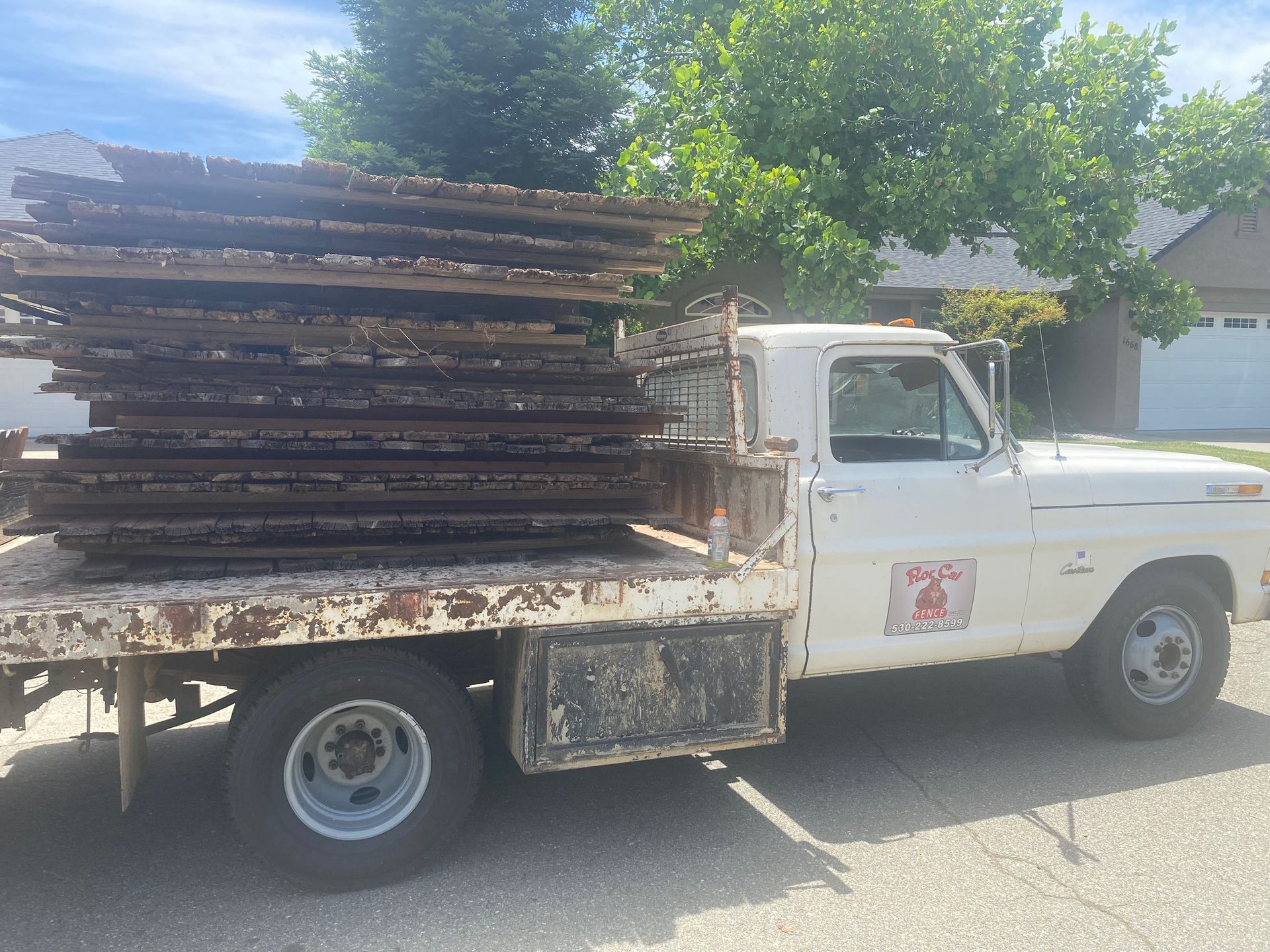 White flatbed truck loaded with stacked wooden planks parked outdoors under trees