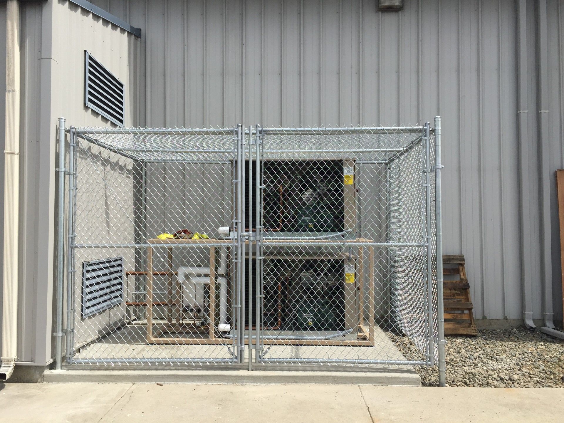 Chain-link fenced utility area beside a gray building, with industrial equipment and vents inside.