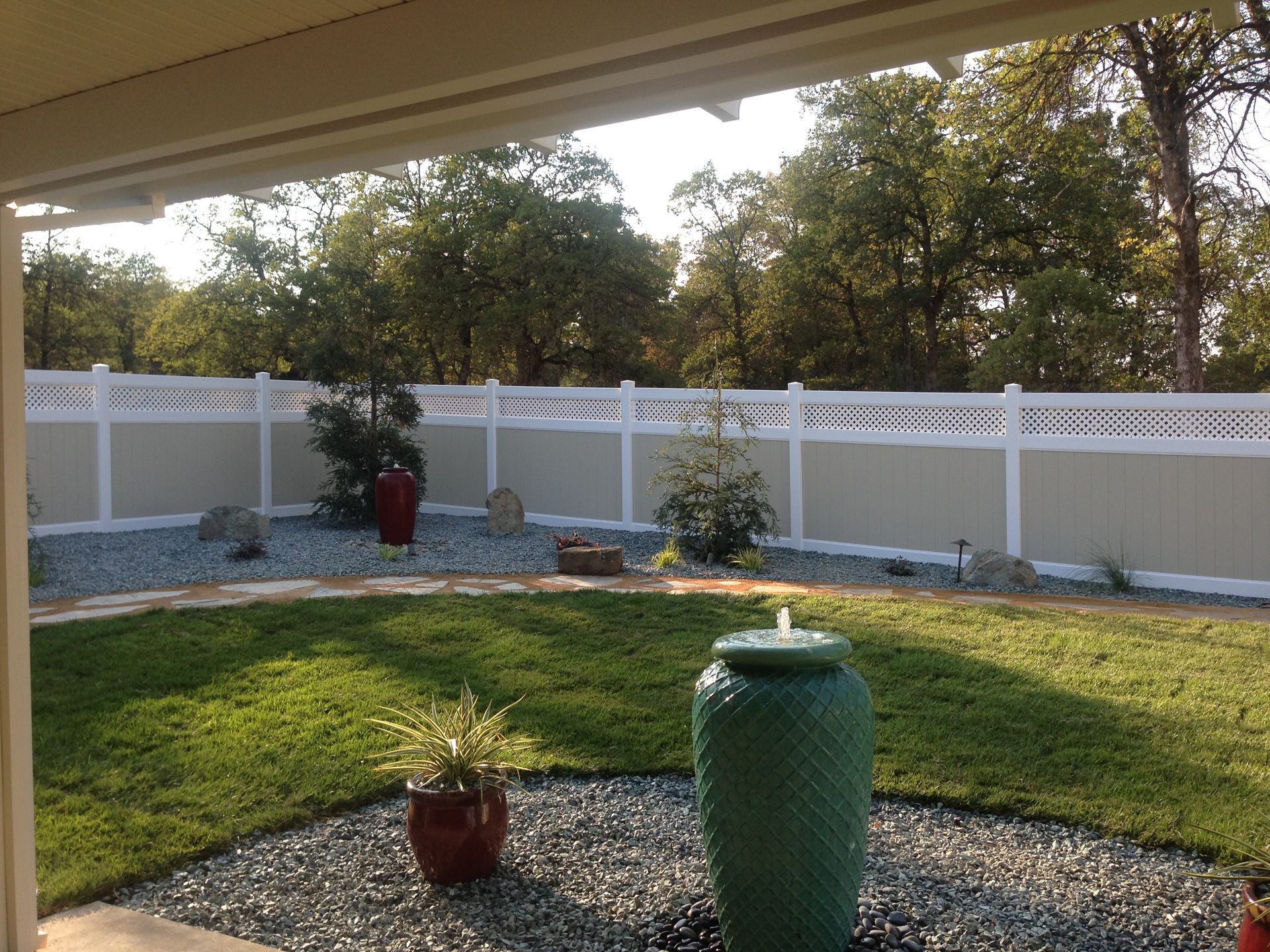 Backyard lawn with a white fence, trees, and a large green ceramic vase in the foreground.
