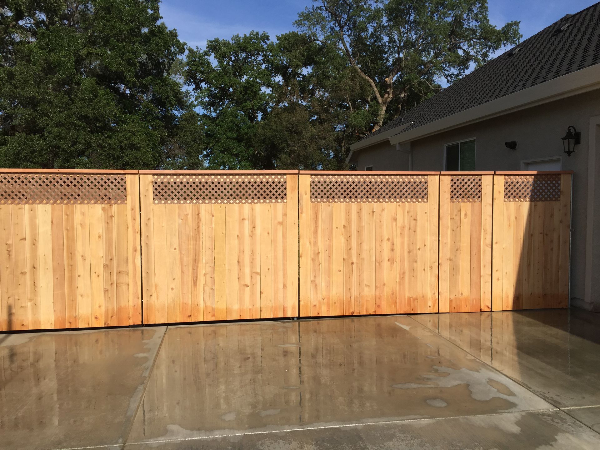 Wooden privacy fence with lattice top beside a house, reflecting on a wet concrete driveway