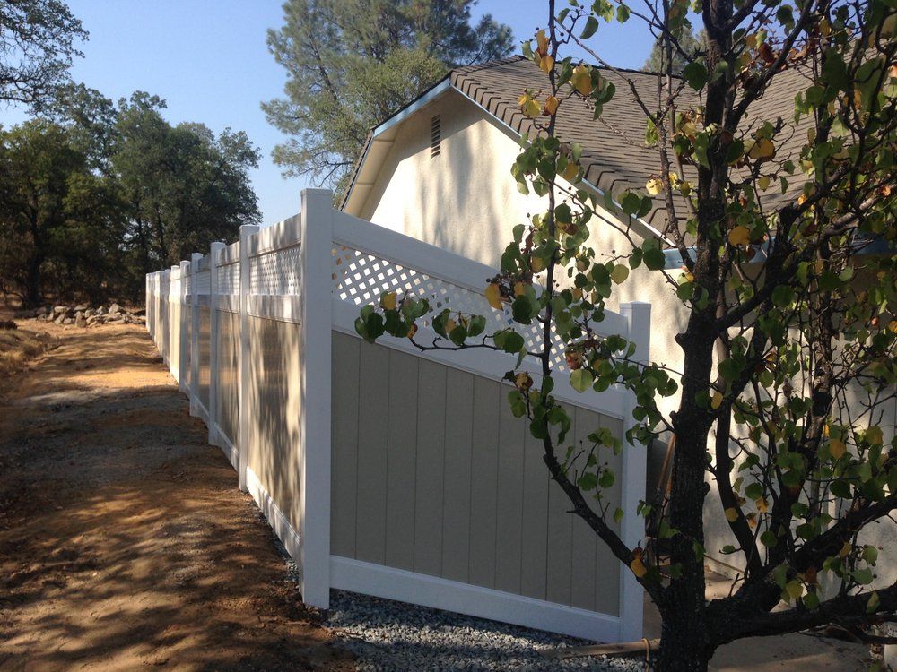 White privacy fence beside a beige house on a wooded dirt driveway in bright sunlight