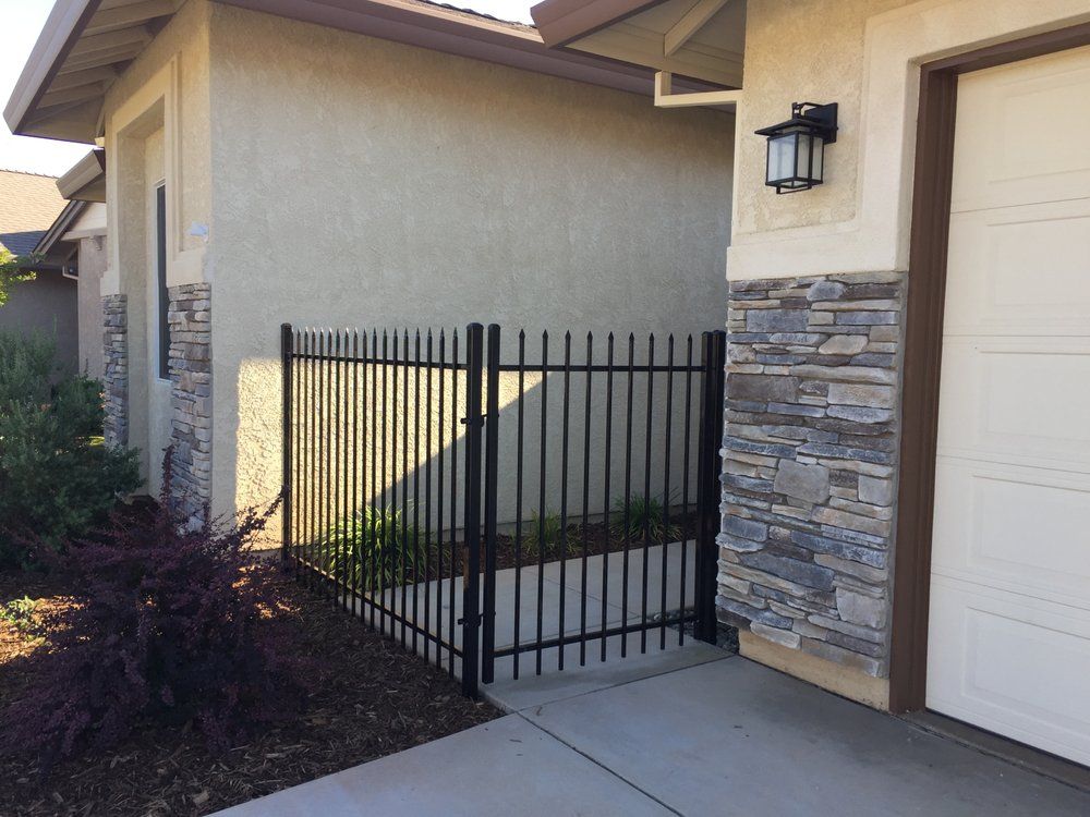 Black metal gate beside a beige house and stone garage wall, with a concrete walkway and landscaping.