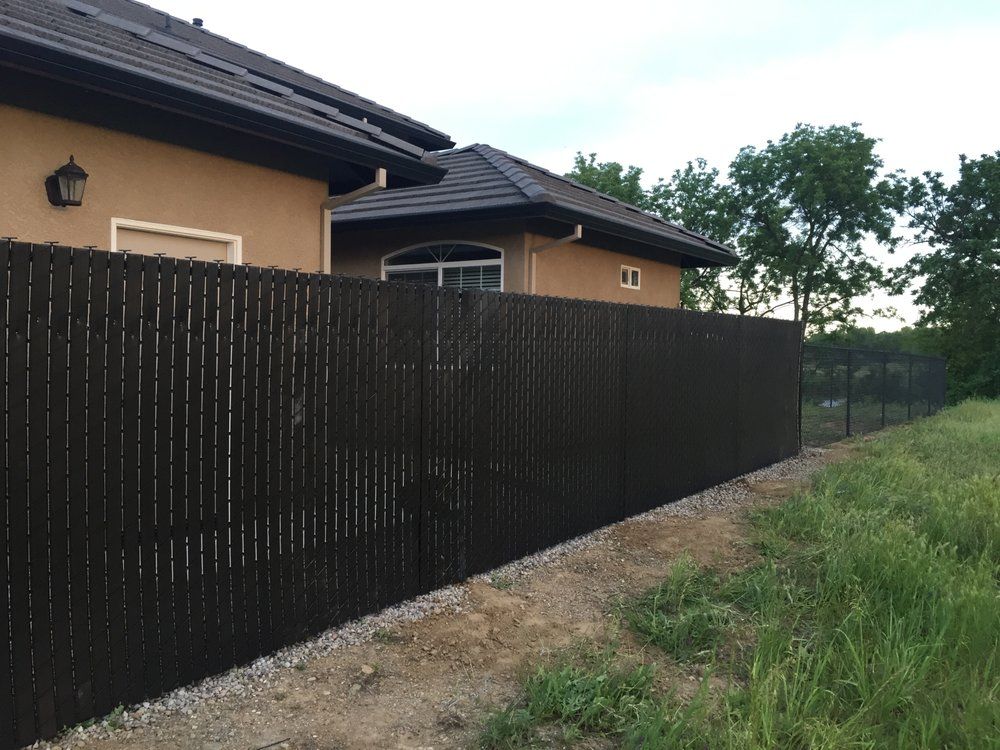 Dark wooden fence along a beige house beside a grassy dirt path and trees