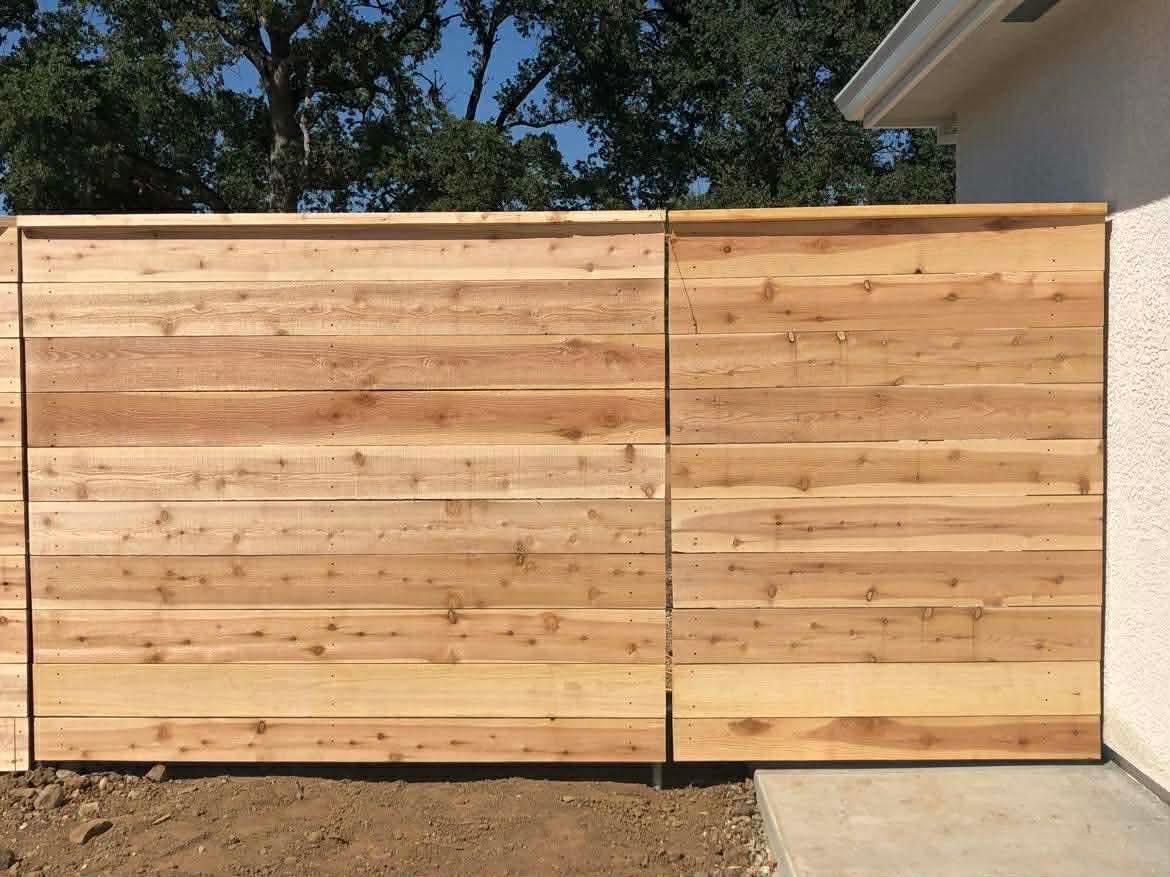 Wooden privacy fence with a gate, bordered by concrete and dirt under a tree canopy