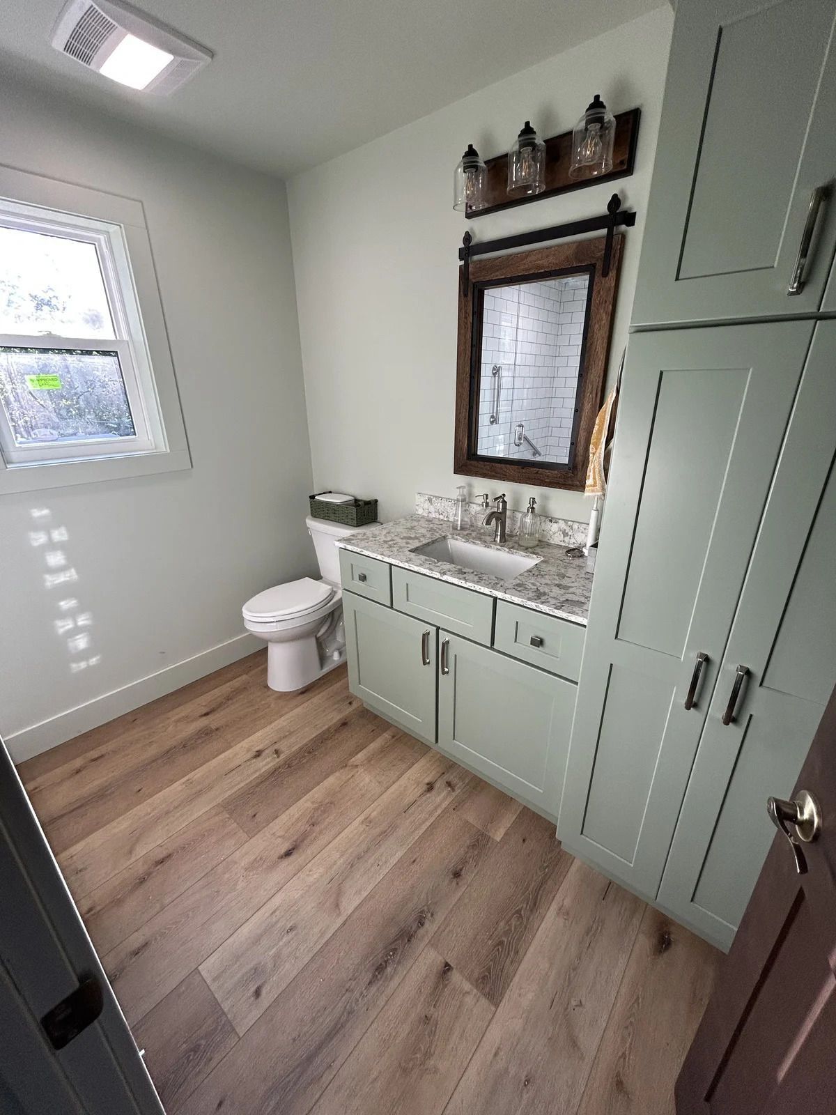 Bathroom with sage green cabinets, light wood floor, white toilet, and window.