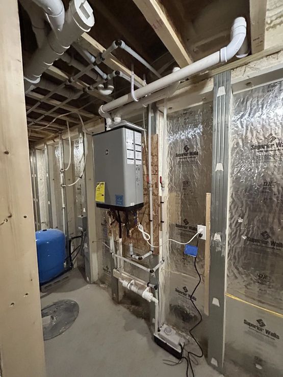 A gray tankless water heater in a basement, with pipes and electrical wiring.