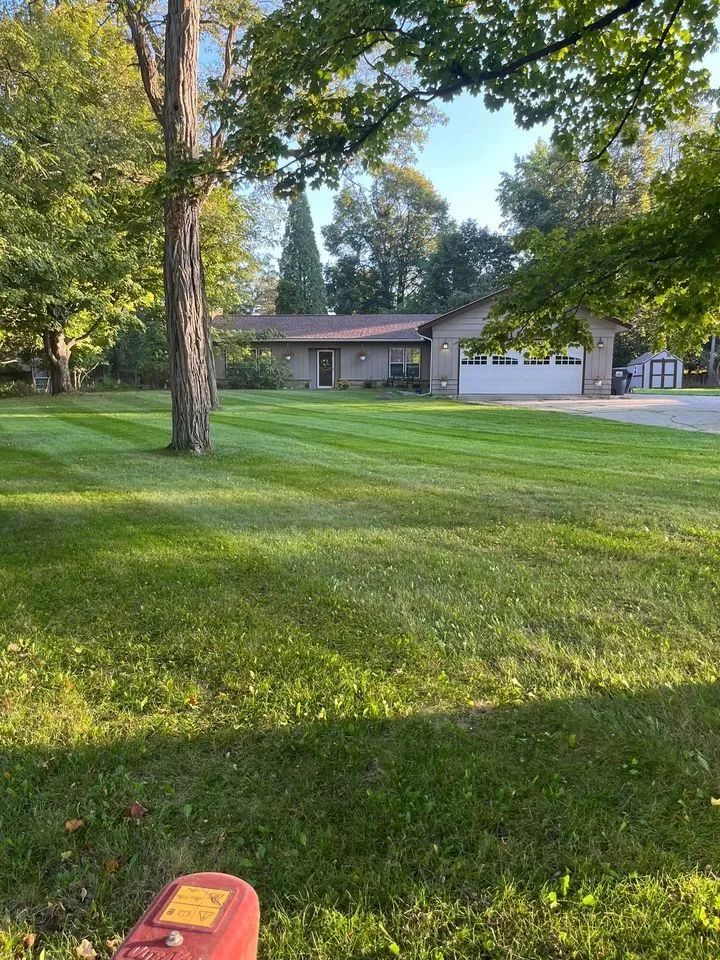 A sprawling lawn with mowed stripes leads to a ranch-style house under a canopy of green trees in the afternoon sun.