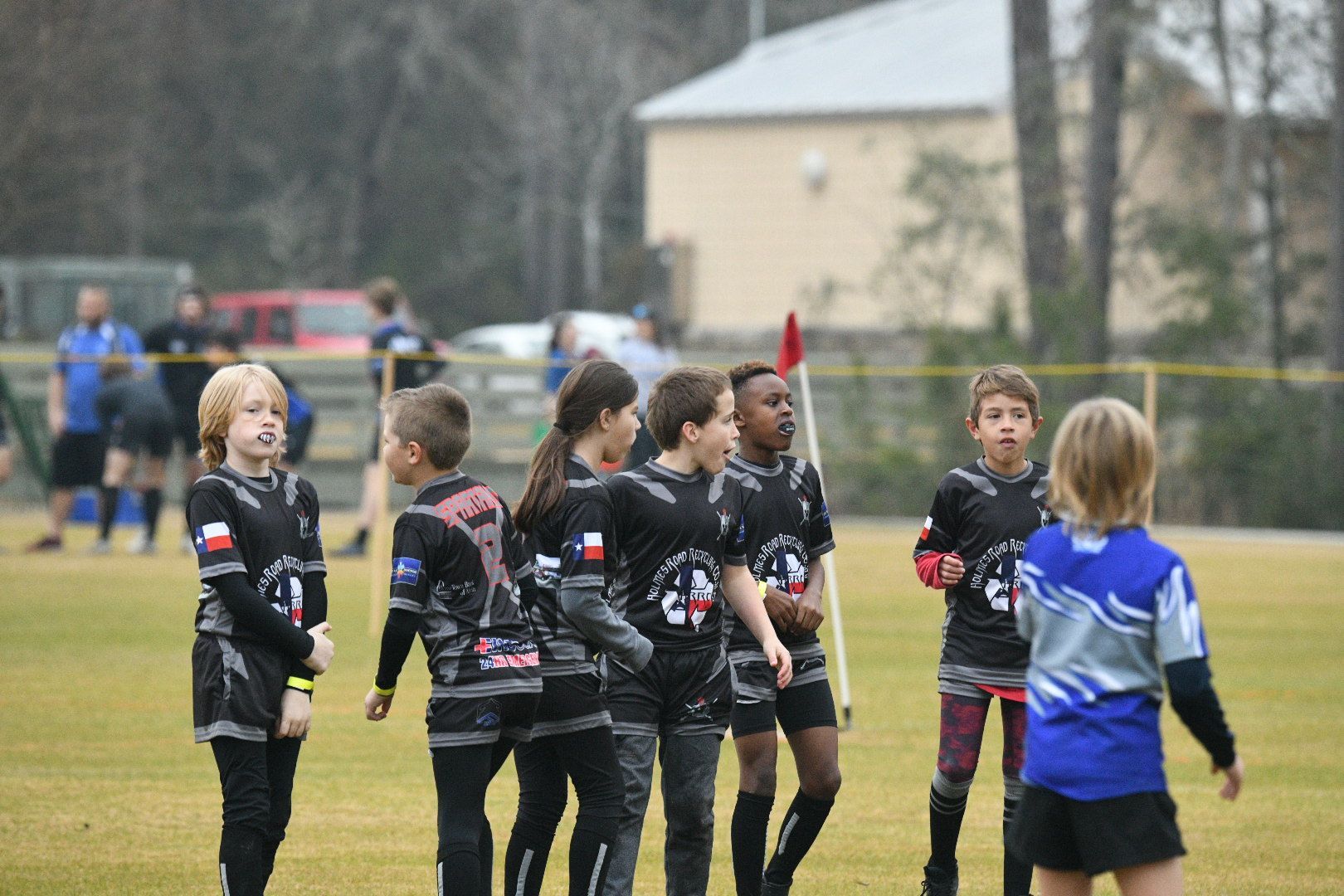 Children in black jerseys huddle on a green field. A blond child in blue stands nearby.