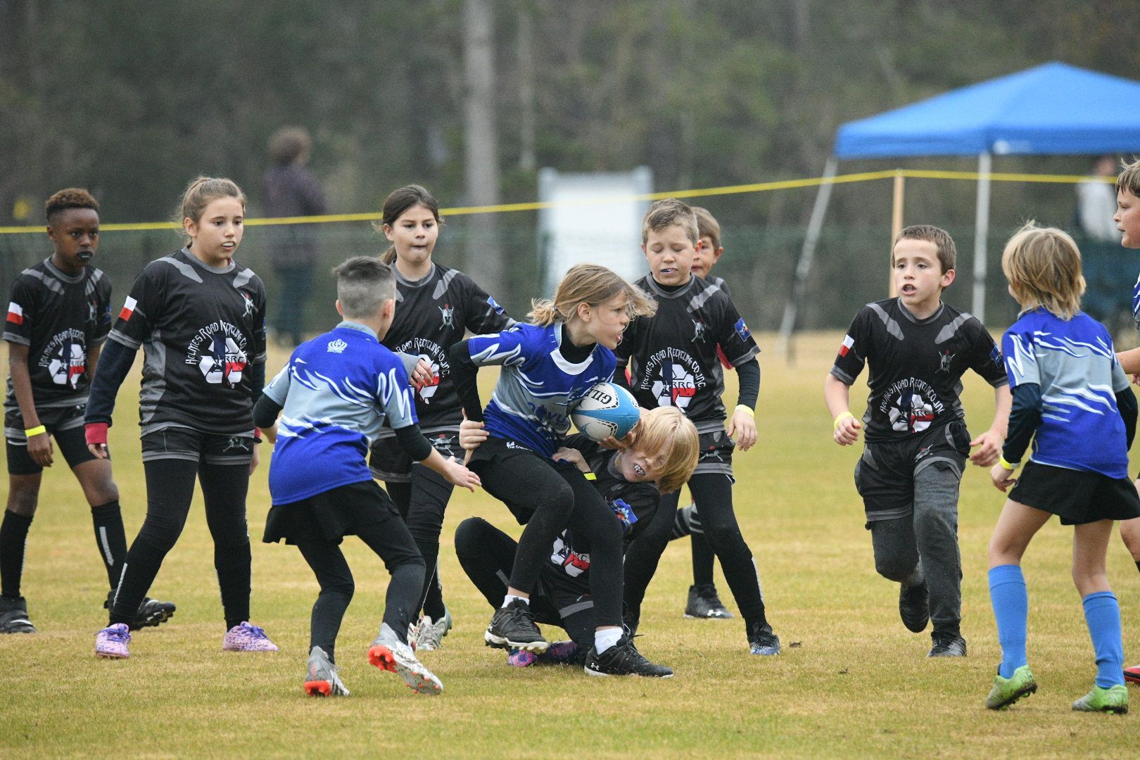 Children playing rugby on a grassy field. Players in black and blue jerseys, one child tackled while holding ball.