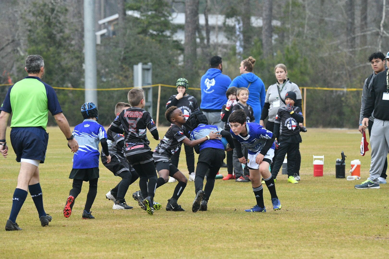 Children in black and blue jerseys playing rugby on a grassy field; referee watches.