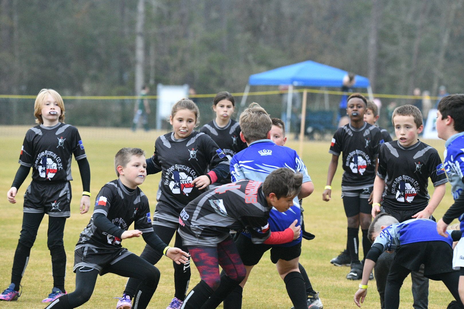 Youth rugby game: Players in black and blue jerseys, tackling on a green field.