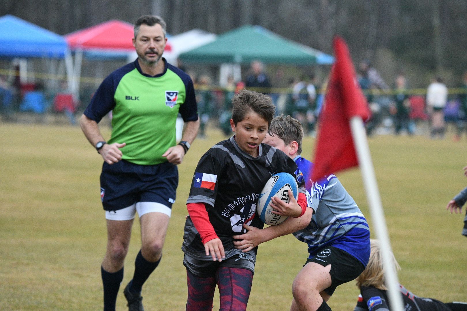 Boy running with rugby ball, being tackled, referee watches field. Outdoors, grassy area.