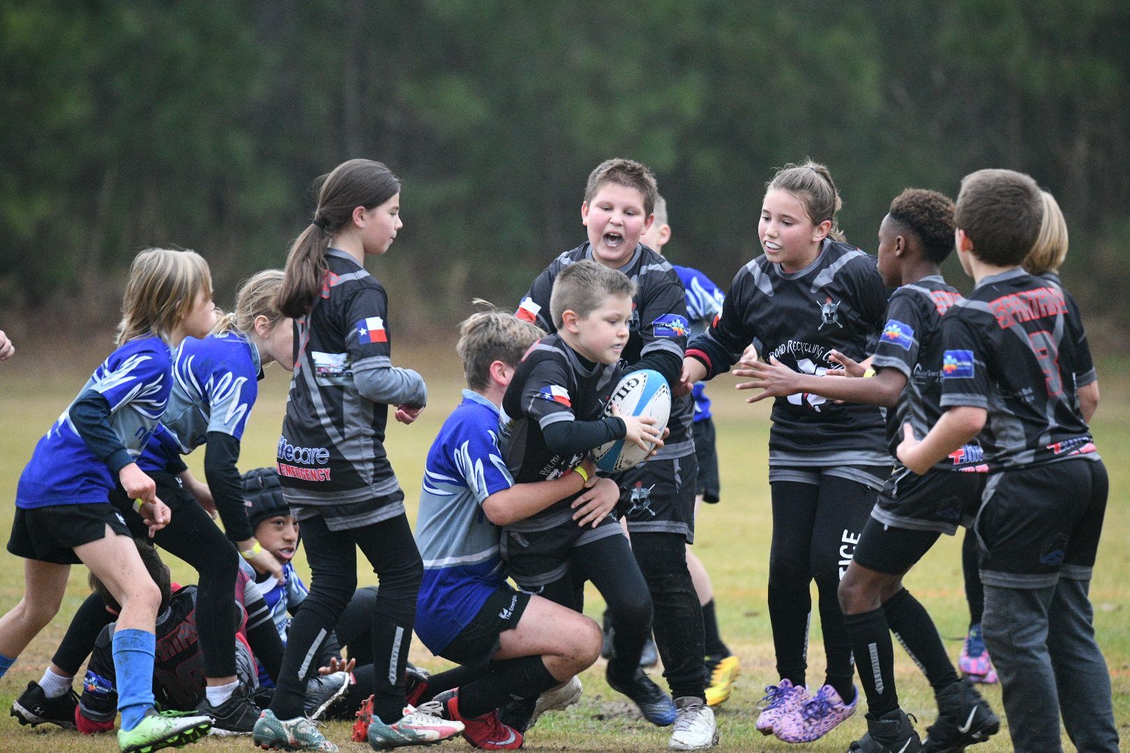 Youth rugby players in a scrum on a field. Some wear black, some blue. Focus on the ball carrier.