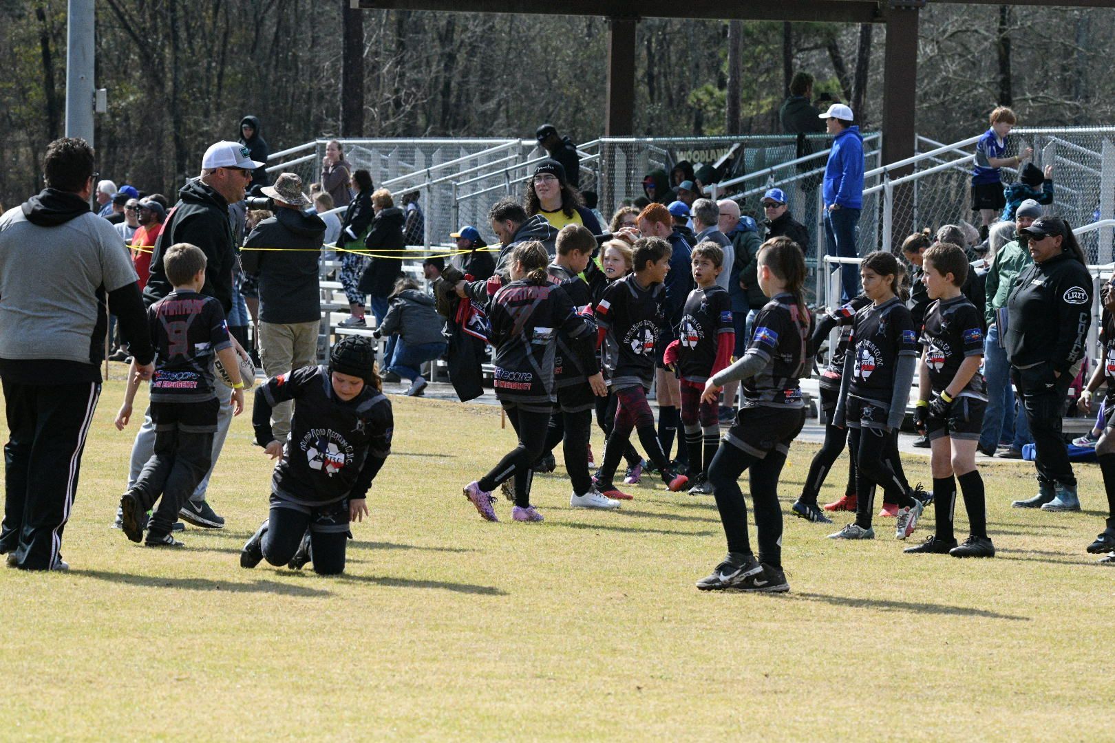 Kids in black and red sports uniforms on a field with adults, likely during a sports event.