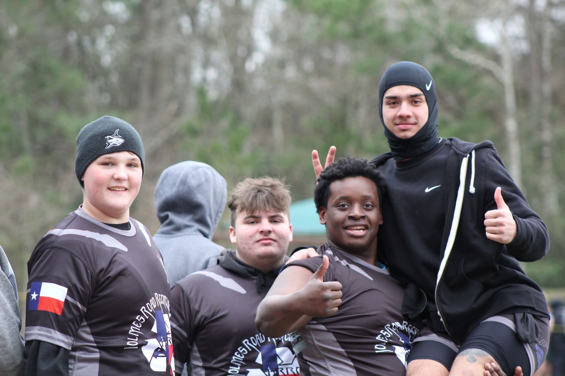 Group of people in black jerseys, smiling, giving thumbs up. Outdoors, trees in background.