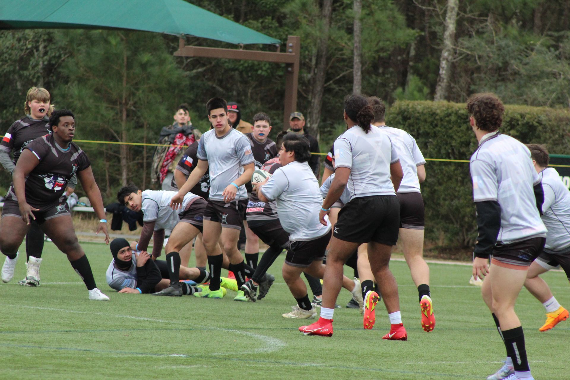Rugby players in grey and black jerseys on a green field. A player runs with the ball, others watch.