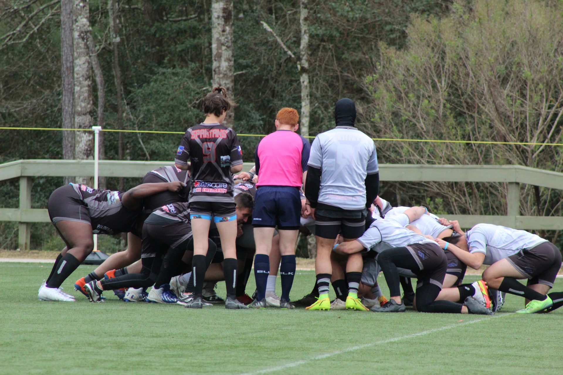 Rugby players in scrimmage, referee in pink shirt, green field, trees in background.