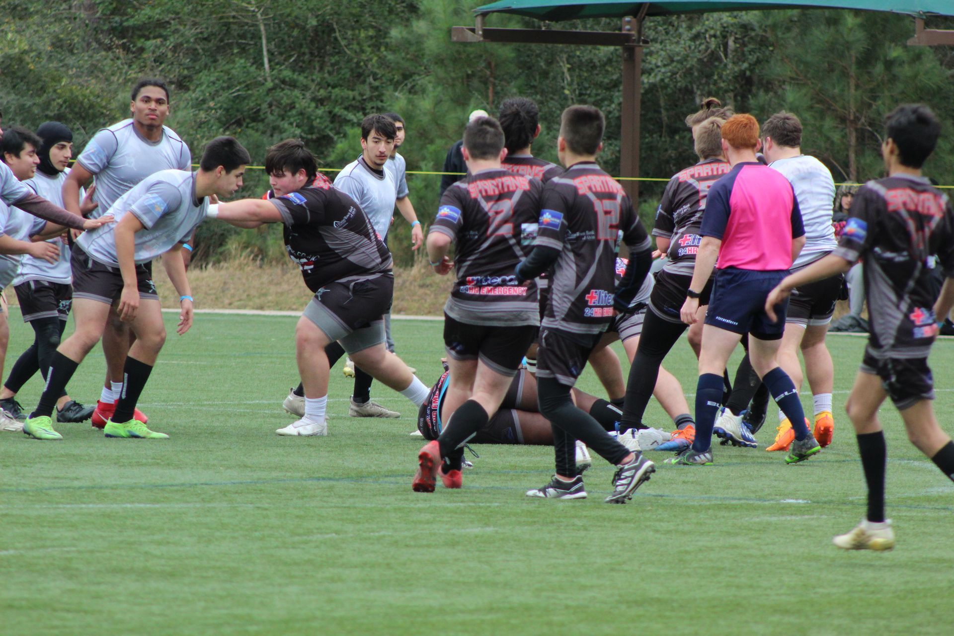 Rugby players on a green field; one player running with the ball, others tackling and supporting; a referee watches.