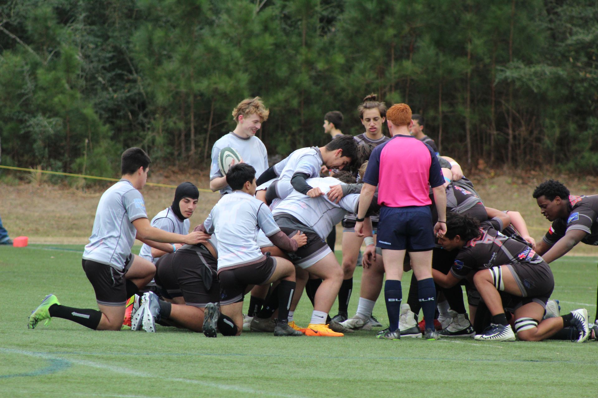 Rugby players in a scrum on a green field. Players in grey and black jerseys huddle together, referee observes.