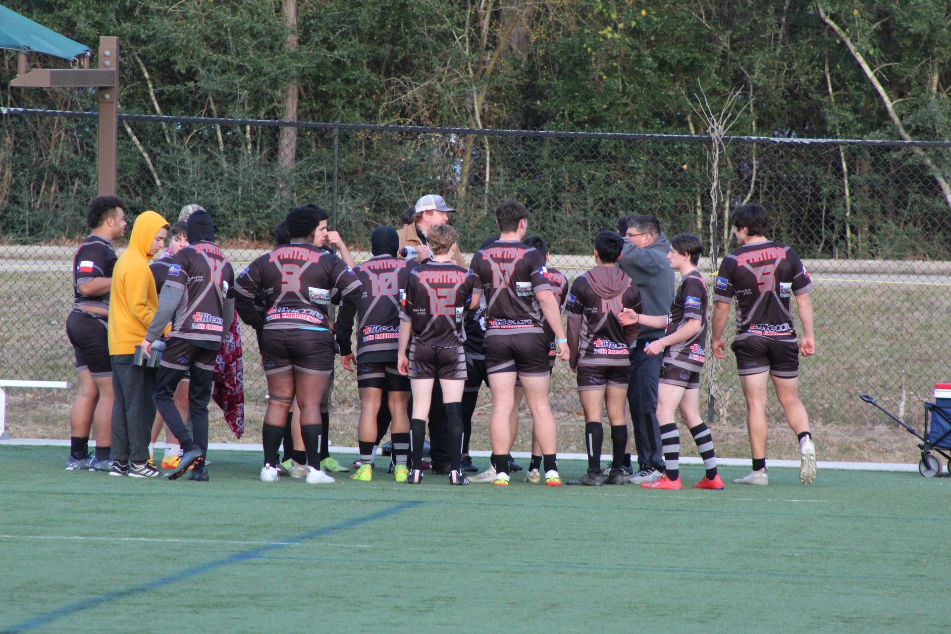 Rugby team huddle on a green field wearing black and pink jerseys, talking with a coach.