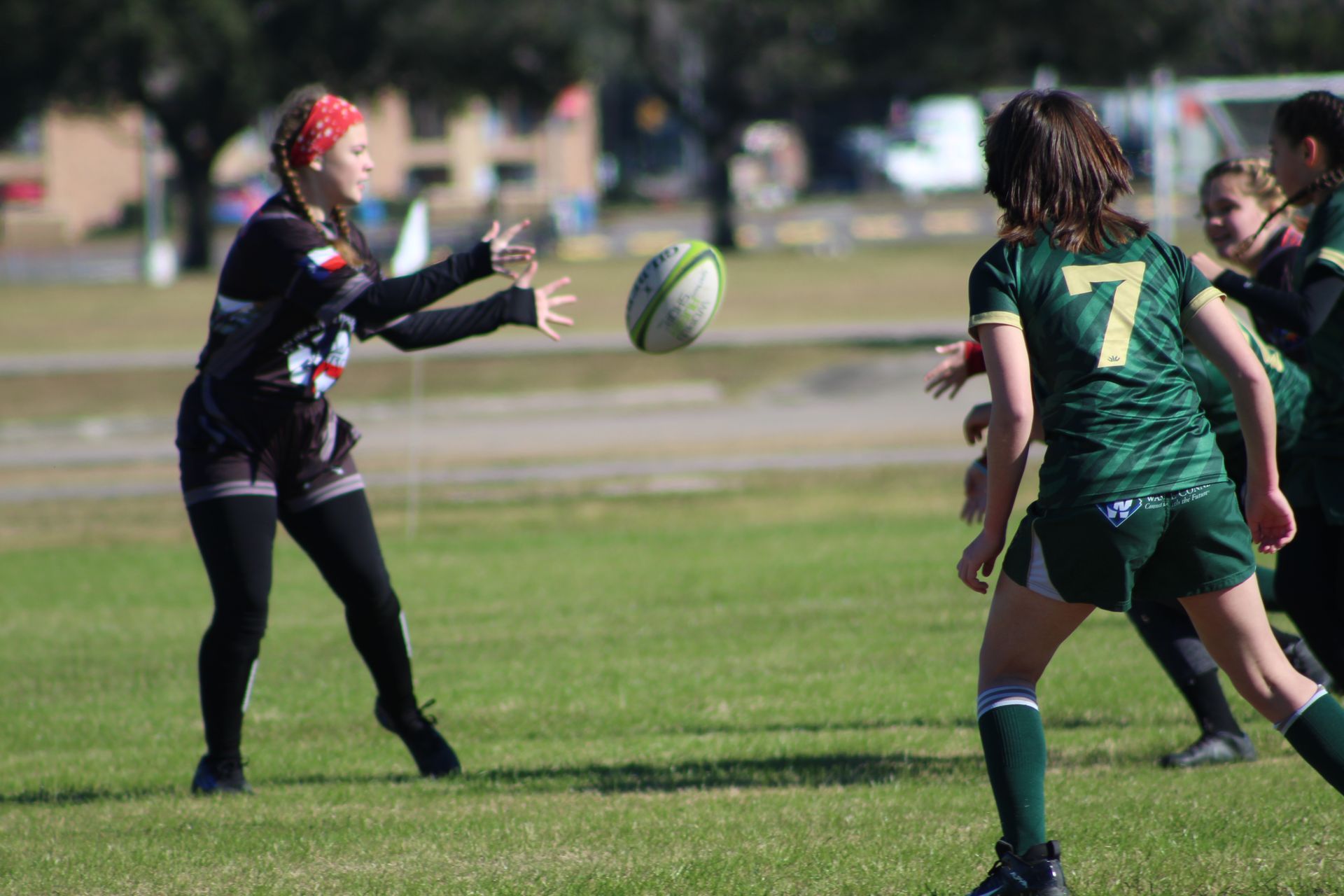Woman in black uniform throws rugby ball to woman in green uniform on grass field.