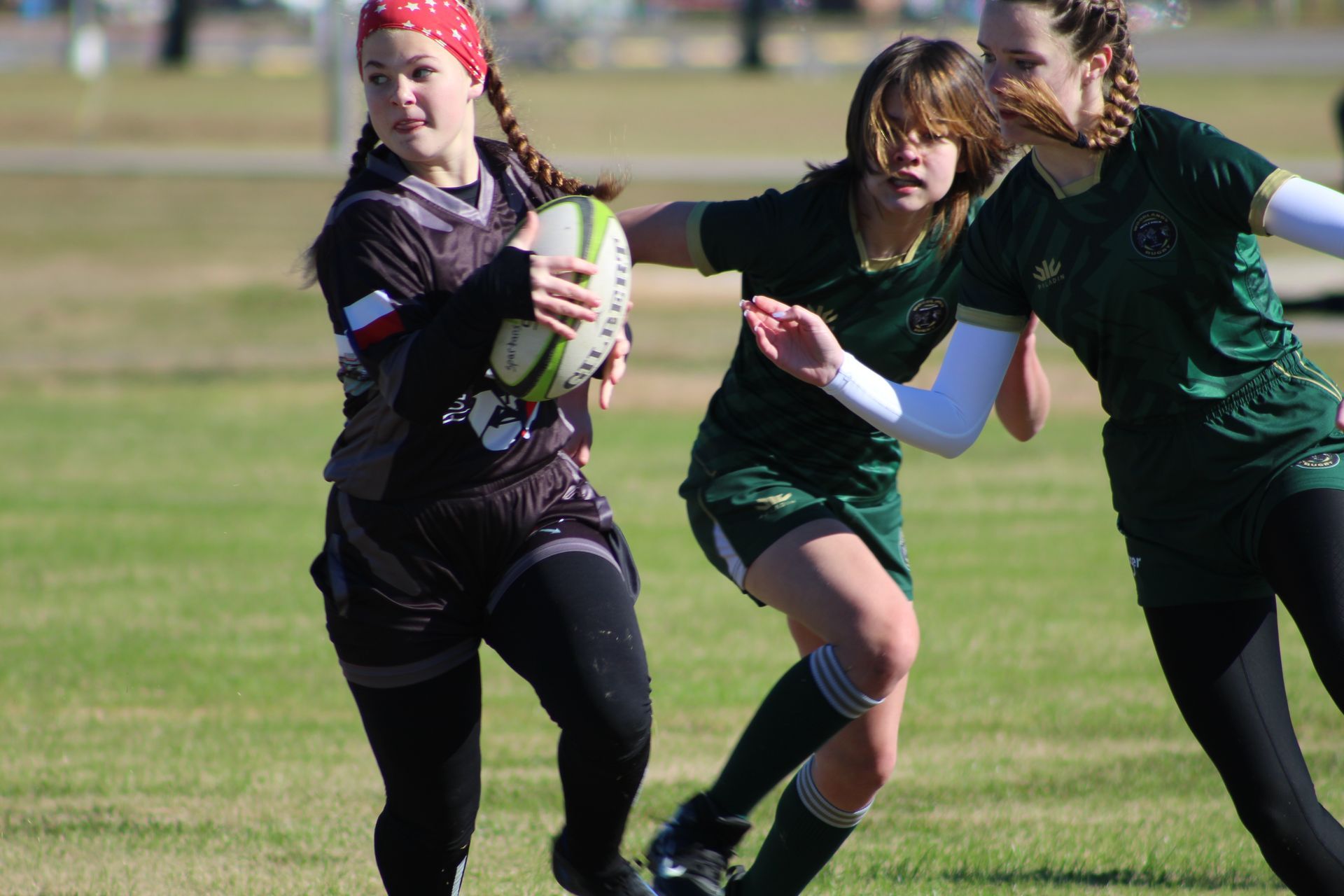 Rugby players on a grassy field. One in black holds a ball, pursued by two in green jerseys.