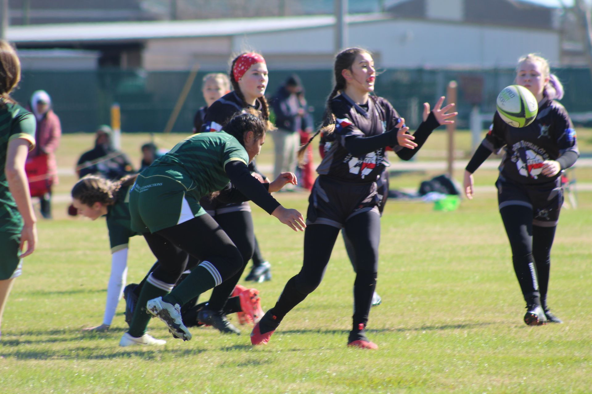 Women's rugby game in progress on a grassy field; player in black prepares to catch the ball.