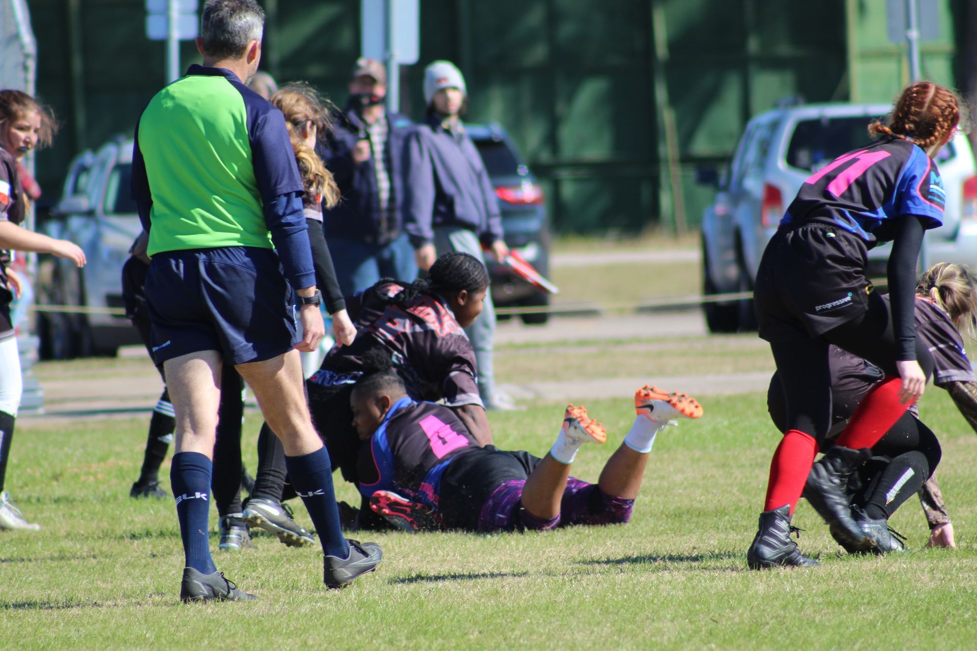 Rugby players in action on a grassy field; one player tackled by another. Referee nearby.