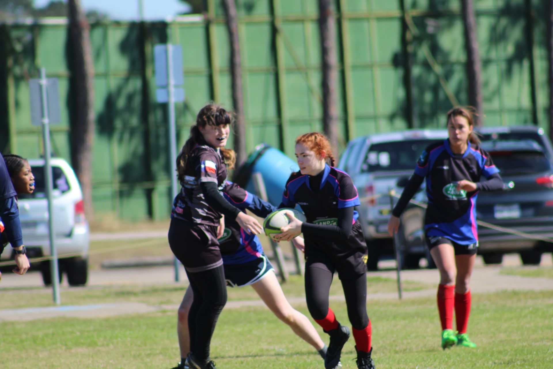 Rugby players in action on a green field. A player in black prepares to pass the ball while another tackles.