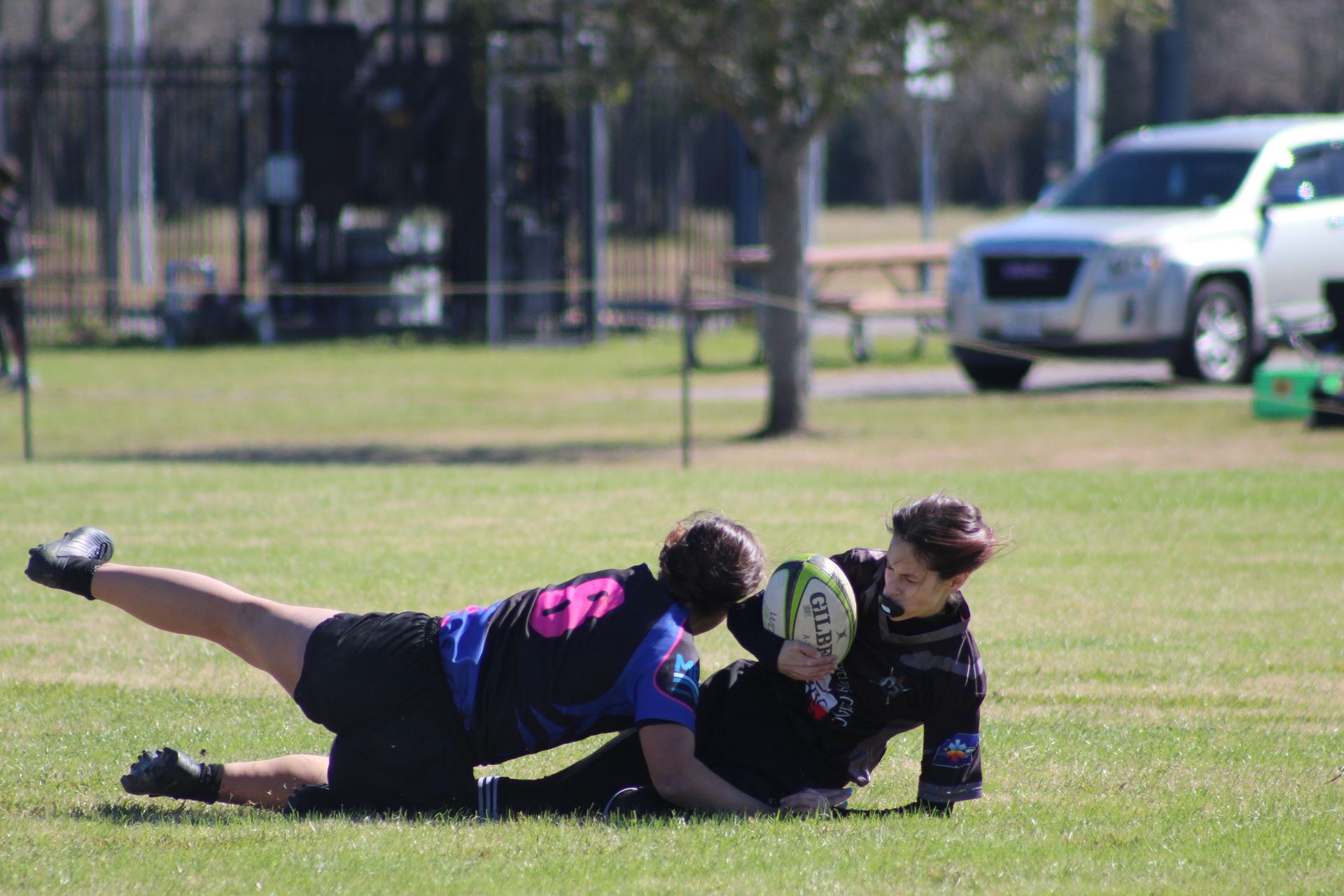 Rugby players on a grassy field; two tackling a player with the ball.