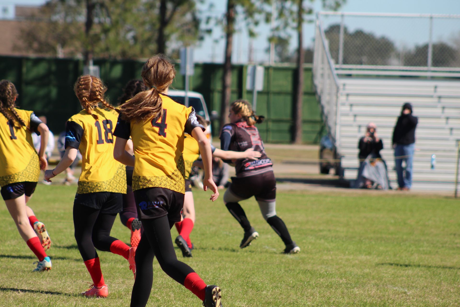 Young women in yellow jerseys playing a sport on a grassy field; bleachers and trees in the background.