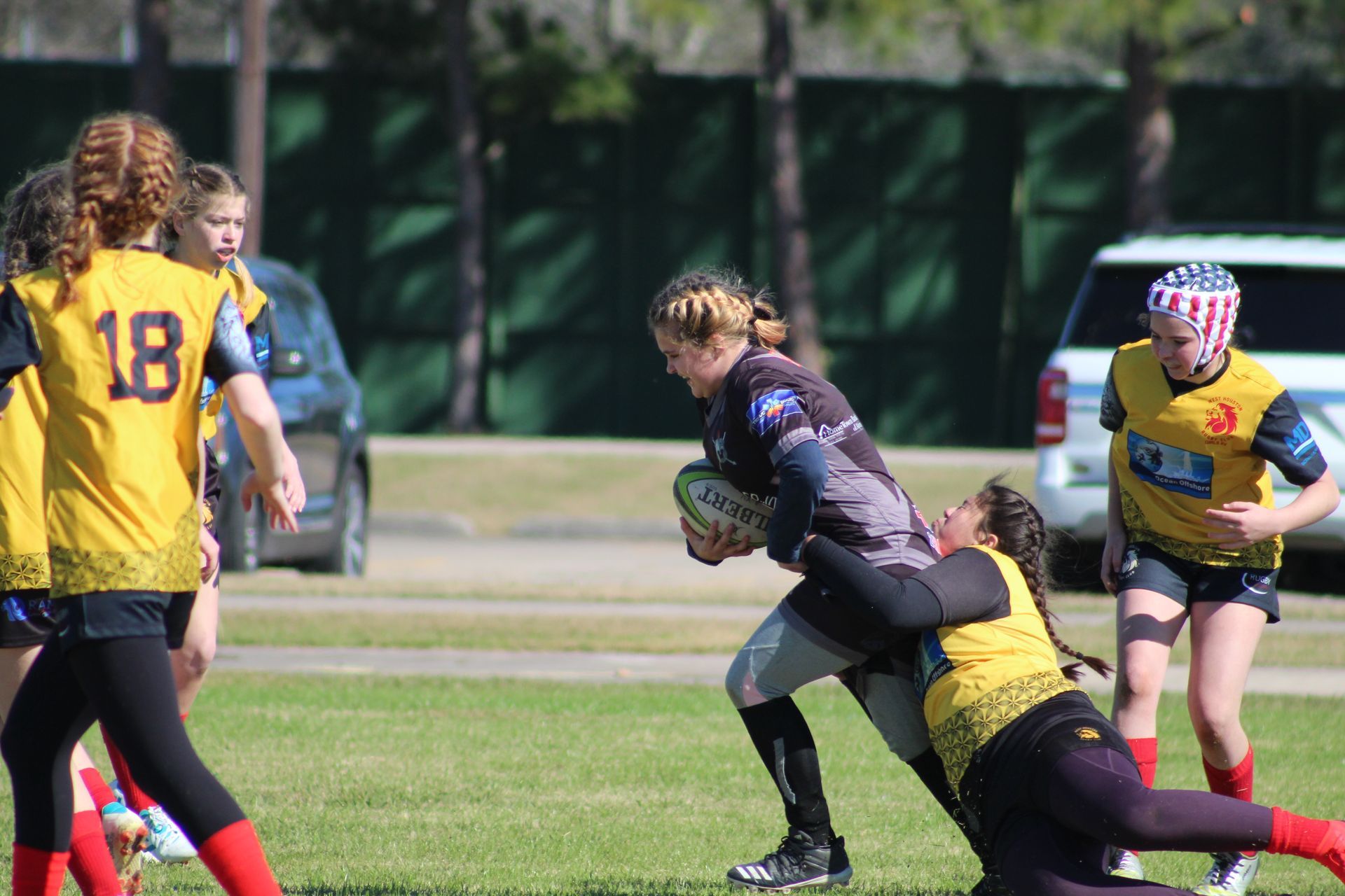 Rugby players on a field. Player in dark uniform runs with ball, tackled by player in yellow. Others watch.