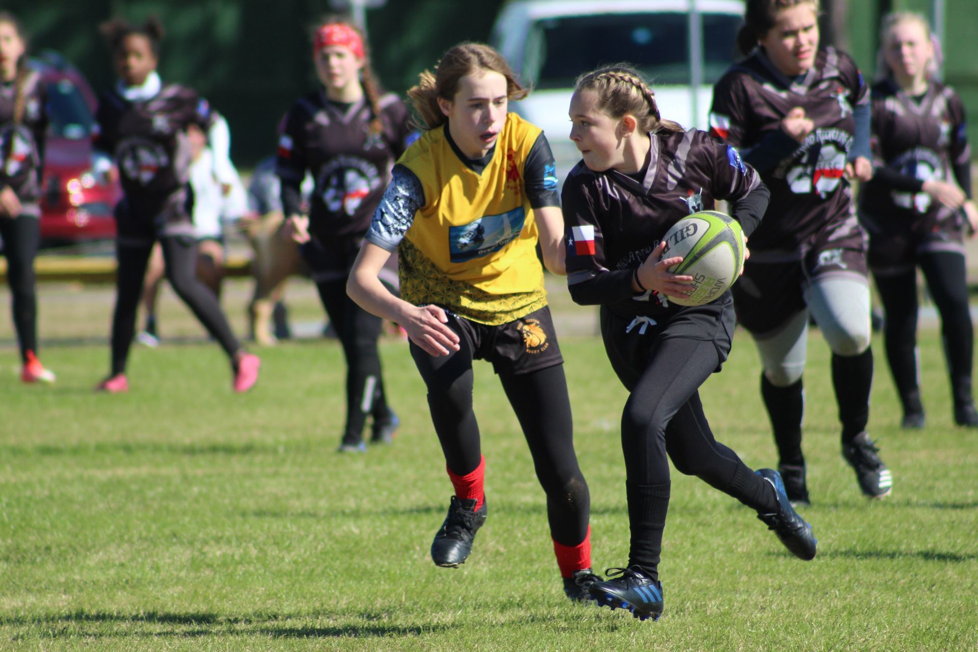 Girls in rugby uniforms playing on a green field. One girl runs with the ball while another tries to tackle.