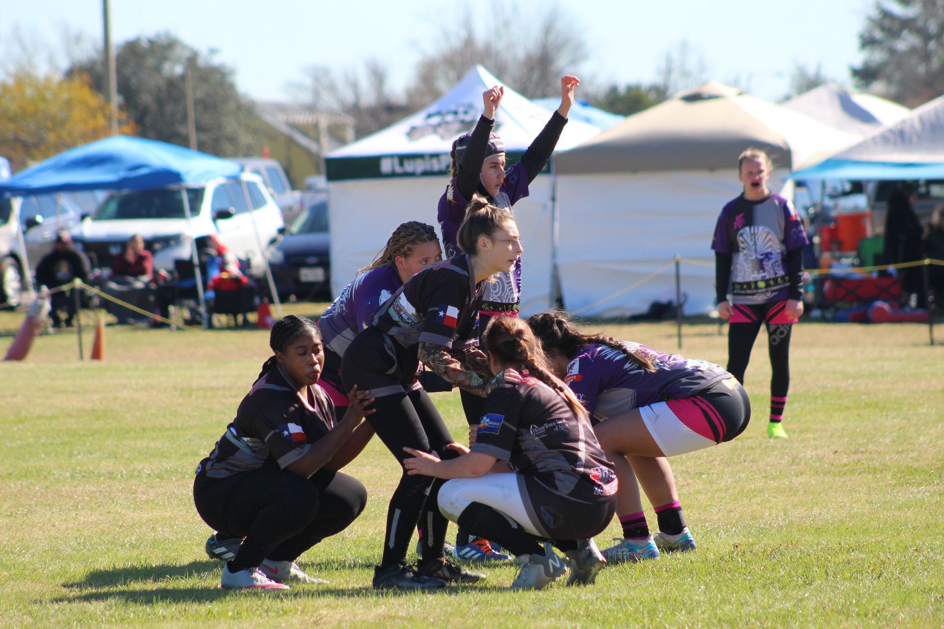 Women's rugby players huddle on a grass field, one raising arms in celebration, others crouched, tents in background.