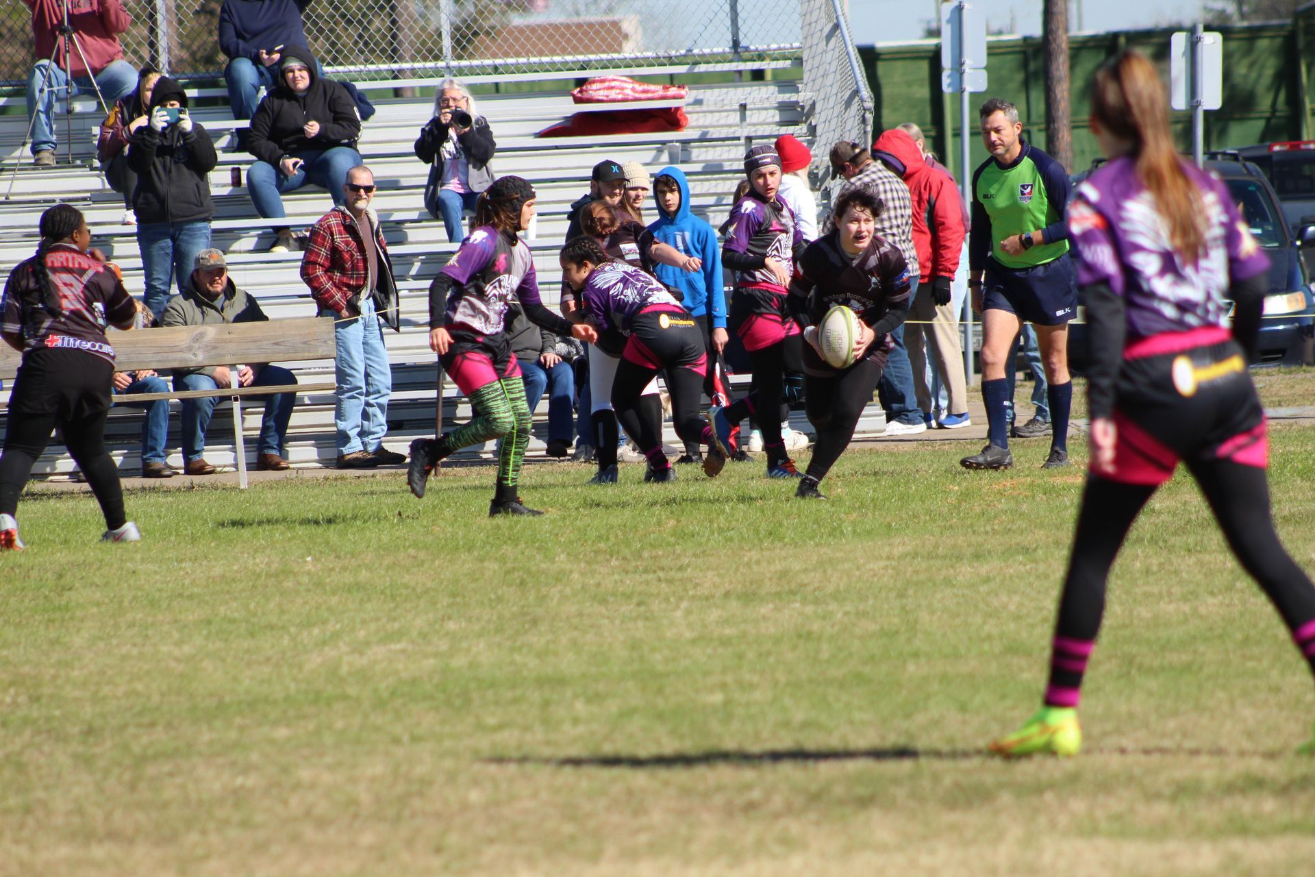 Rugby players in black, purple and pink uniforms playing on a grassy field. Spectators watch from the sidelines.