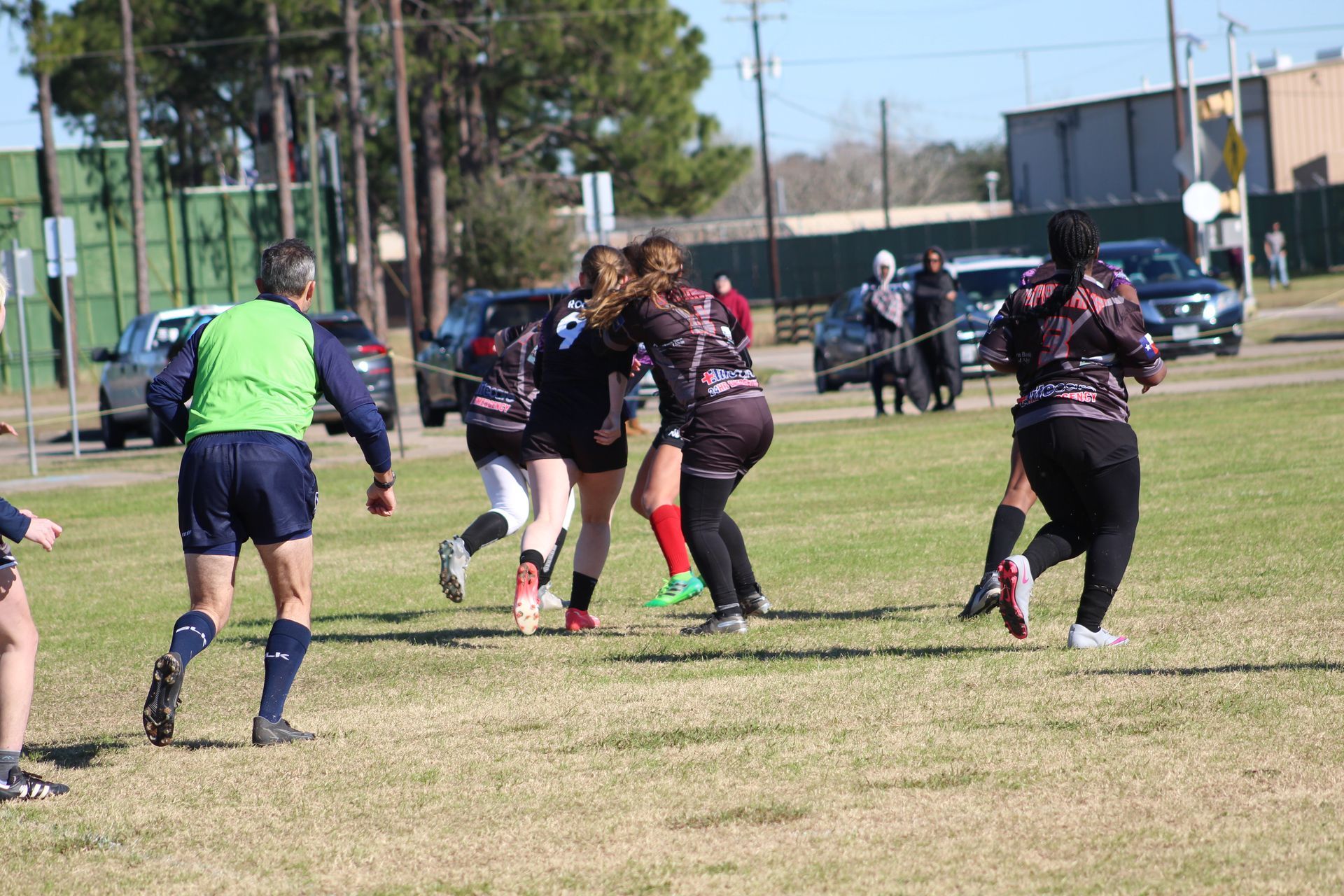 Rugby game in progress; players in black and maroon uniforms running on a grassy field, referee in green.