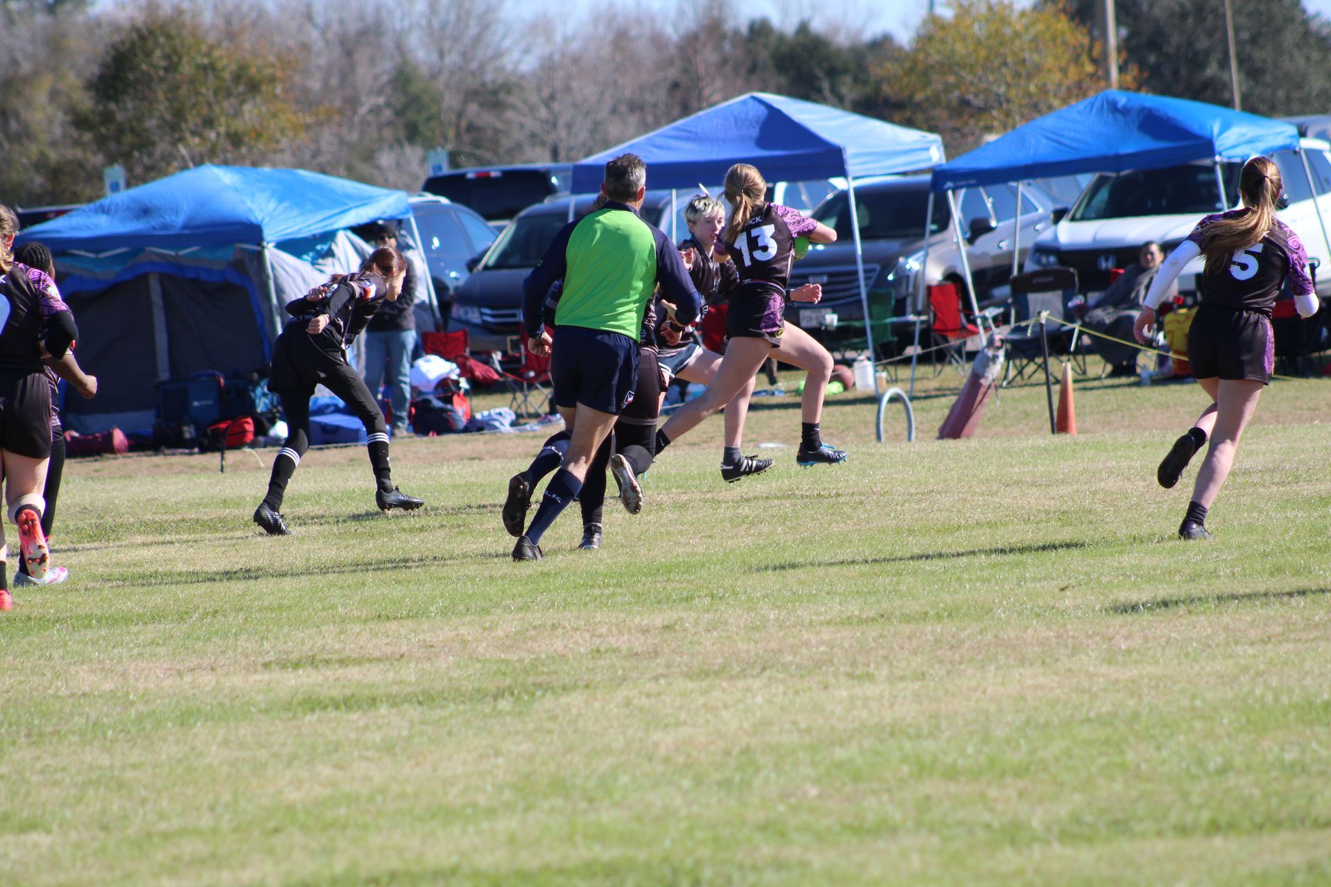 Female rugby players in action on a grassy field, blue tents in background. Referee watches the play.