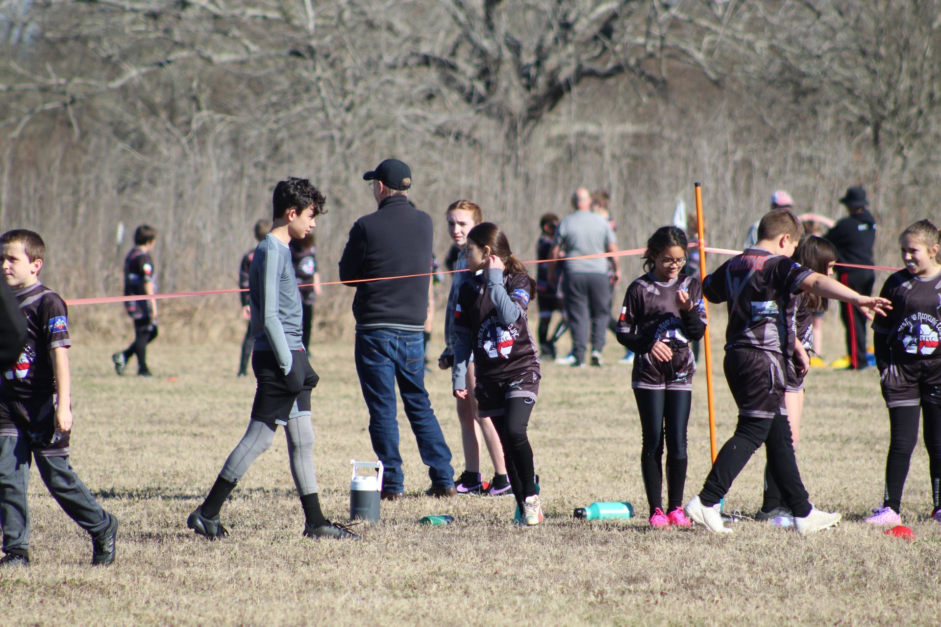 Children in athletic wear at a cross-country event, some near a fence, others running on a field.